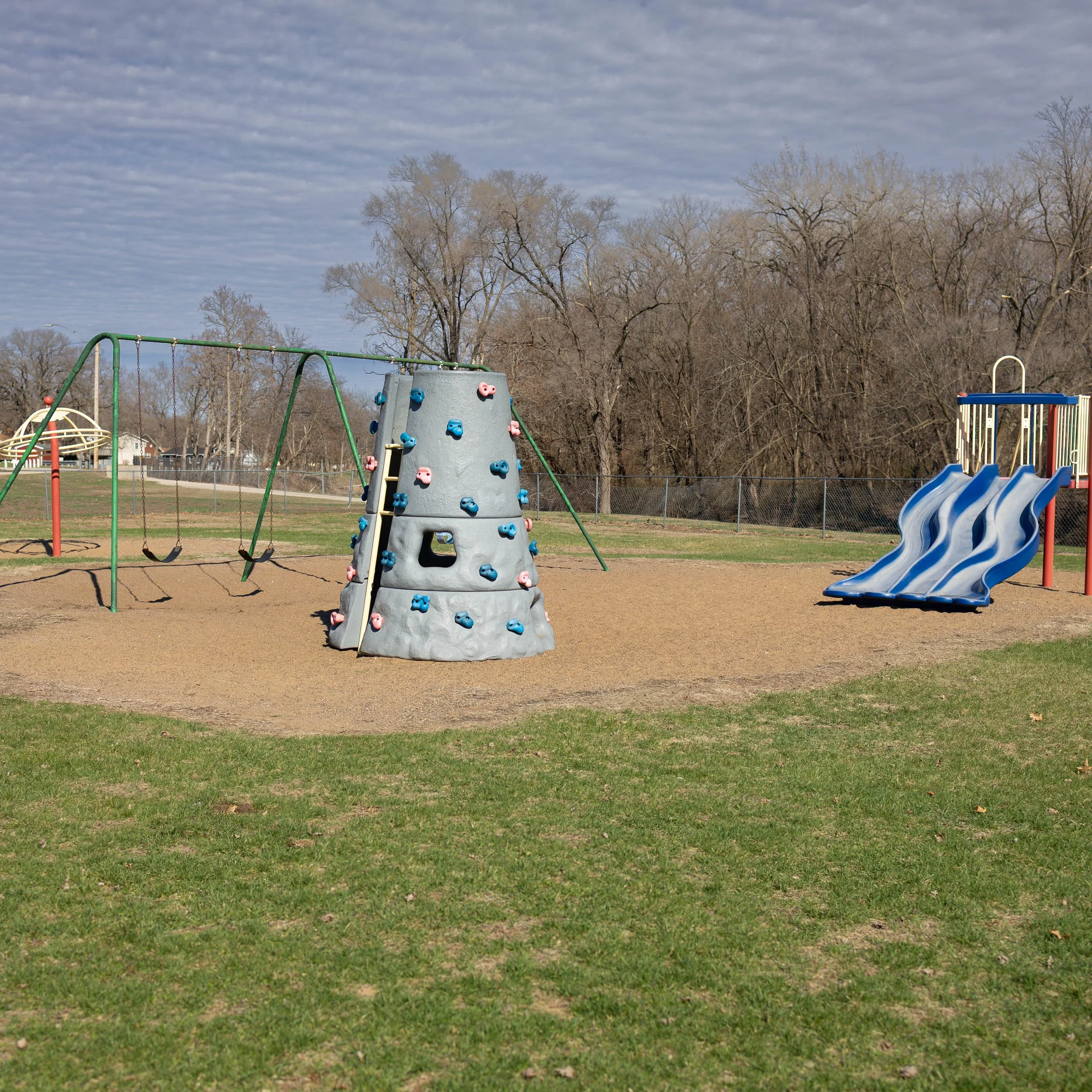 Empty playground with swings, climbing rock wall, and slides, fenced in with leafless trees in the background on a partly cloudy day.
