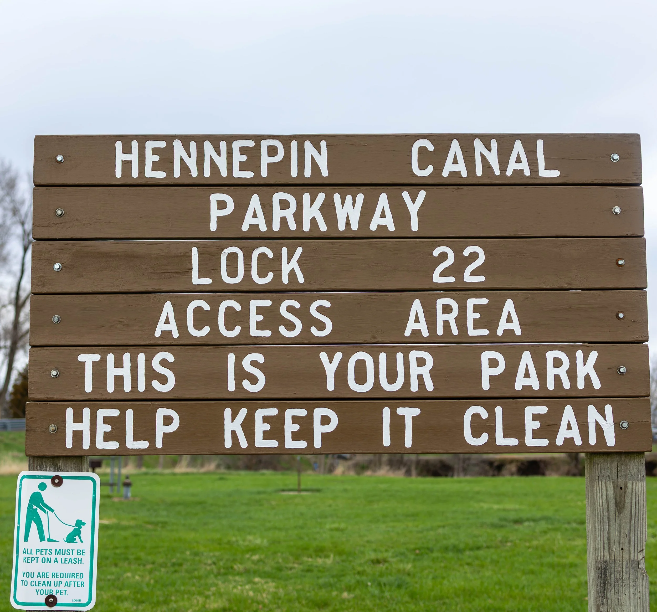 A wooden park sign with white lettering indicating directions and rules for a park, including Hennepin Canal Parkway Lock 22 Access area, and a separate green and white sign below requiring pets to be on a leash and owners to clean up after them.