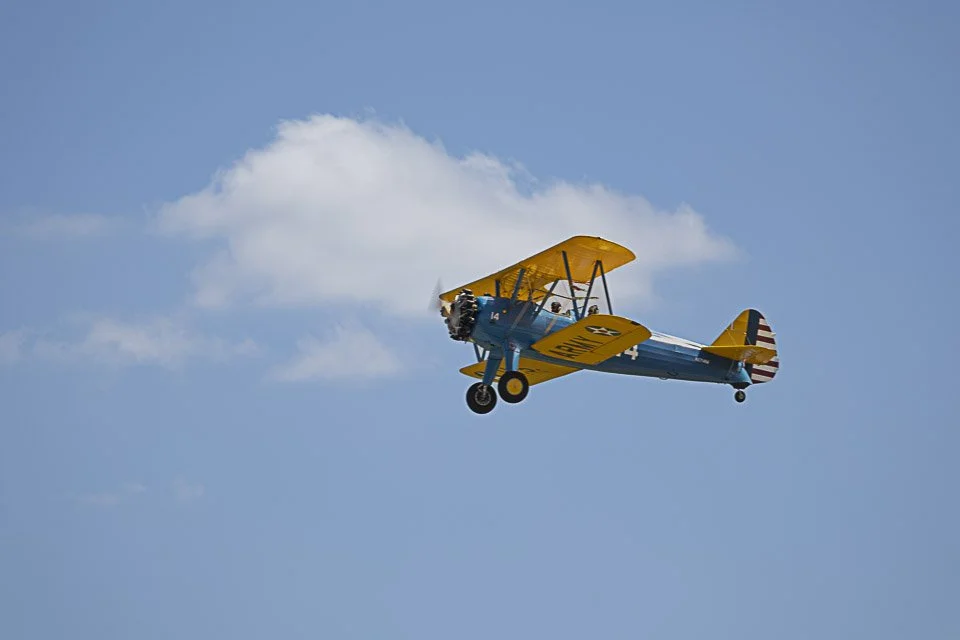 A vintage yellow and blue biplane flying in a clear blue sky with a few white clouds.