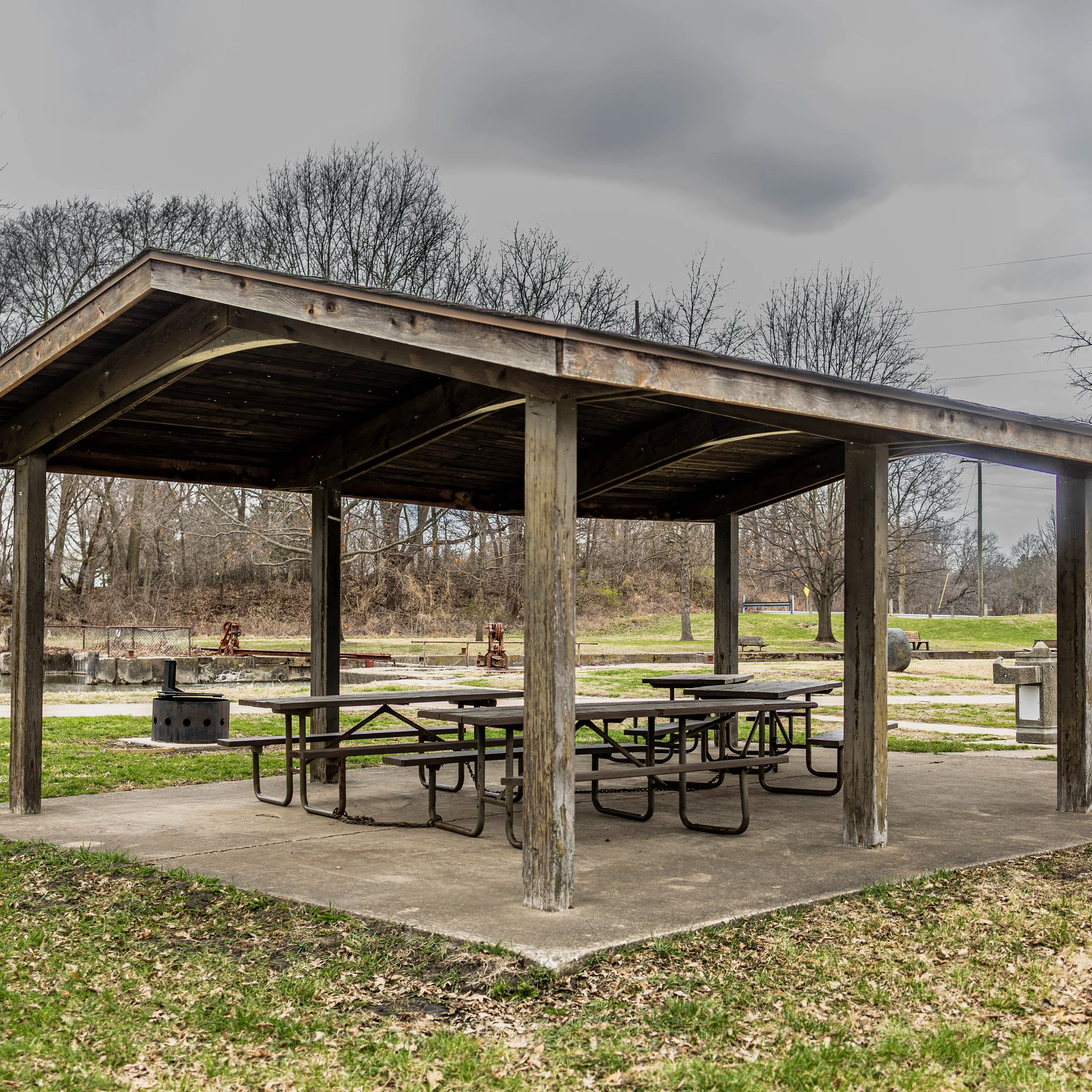 A park pavilion with a wooden roof and metal picnic tables underneath, surrounded by grassy area and leafless trees, on a cloudy day.