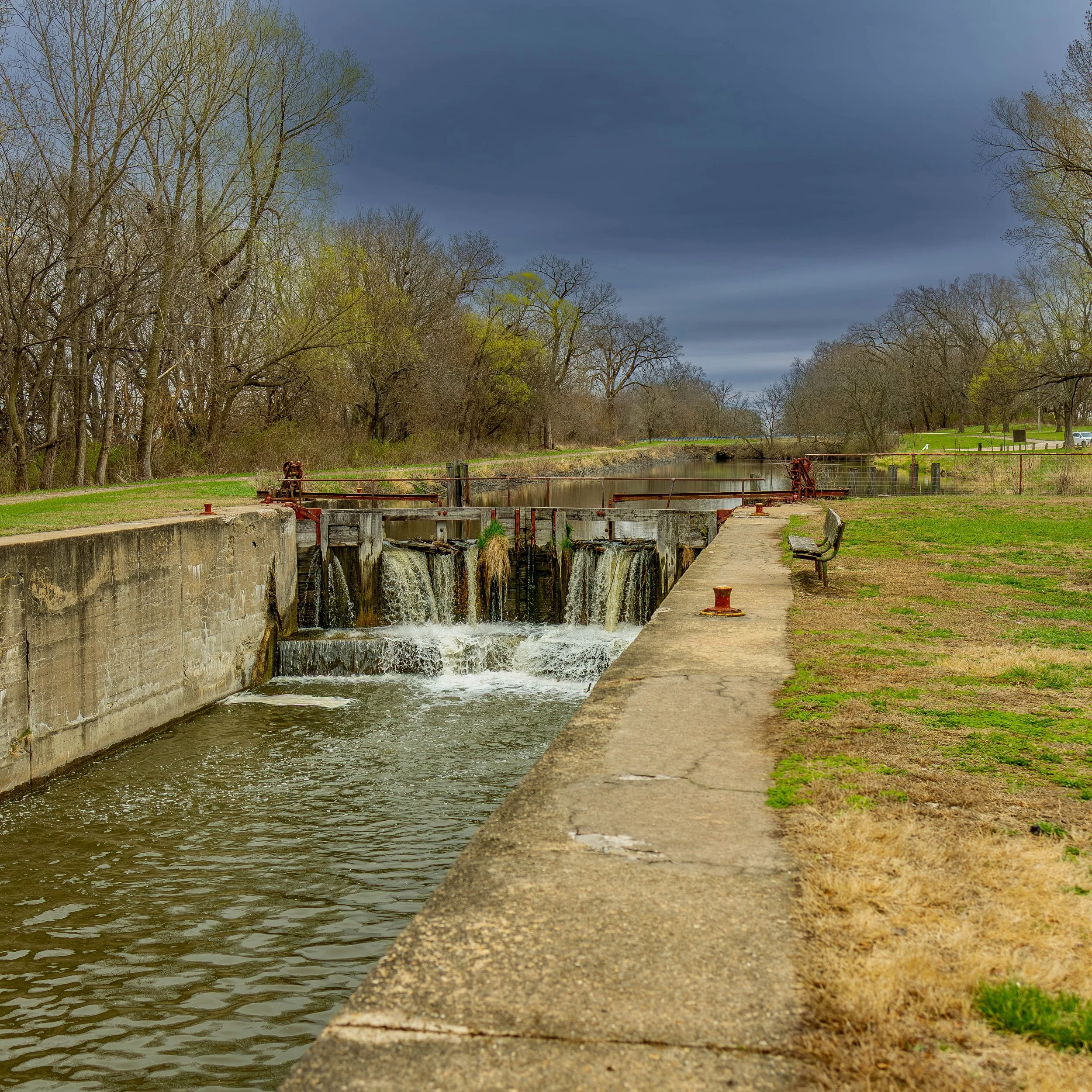 A small dam with water flowing over it, adjacent to a concrete walkway, with trees and grassy areas on both sides and a cloudy sky overhead.
