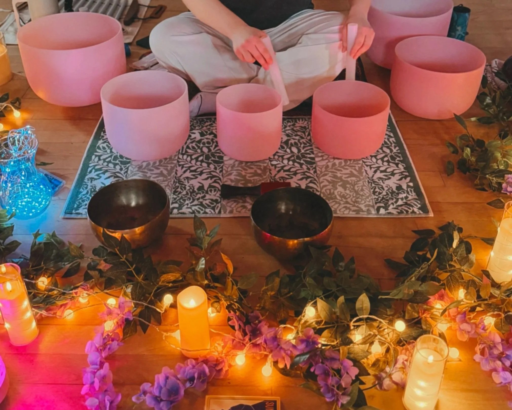 Person sitting on floor surrounded by pink singing bowls, decorative candles, string lights, greenery, and flowers, preparing for sound therapy session.