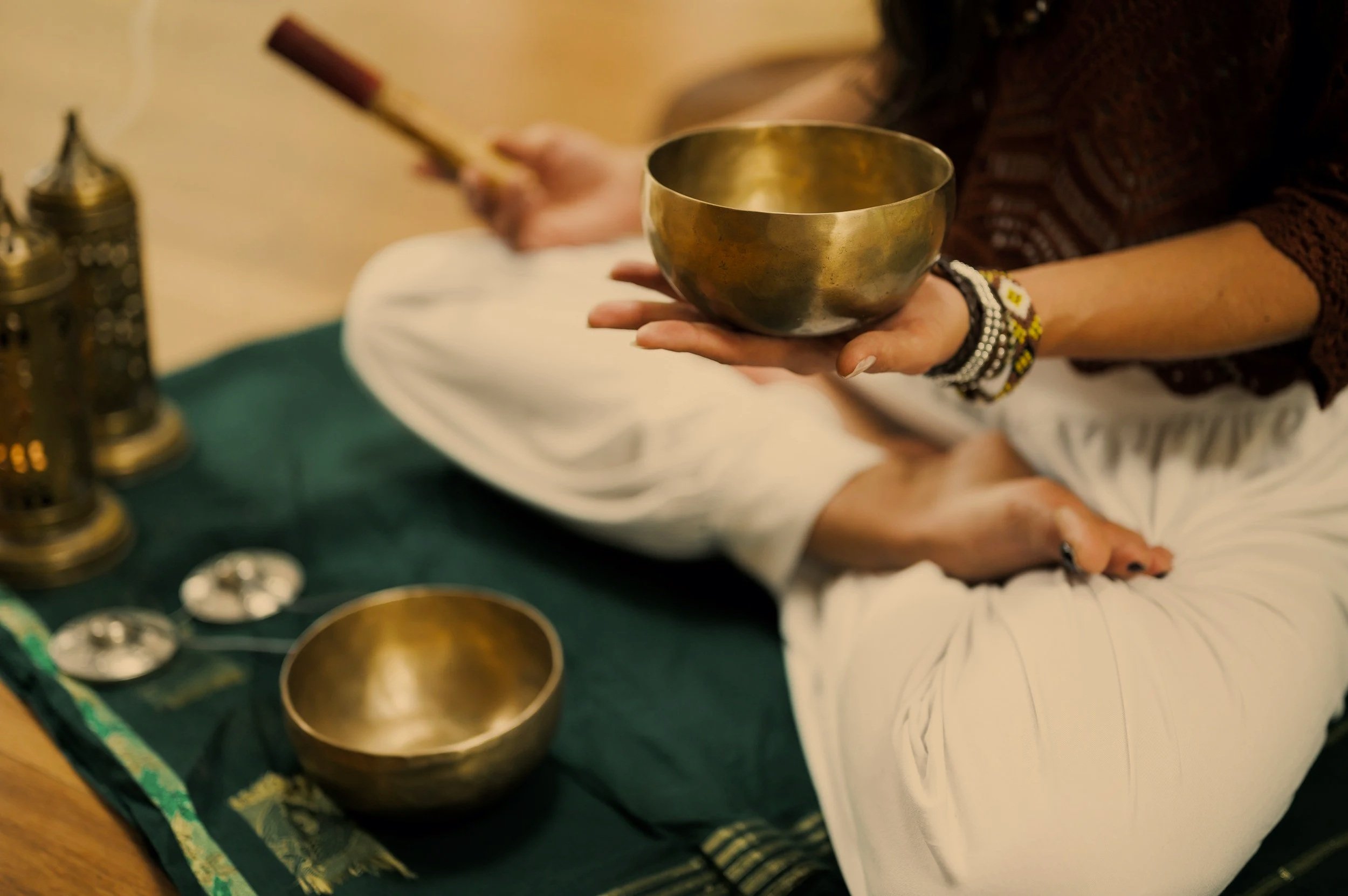 Person holding a singing bowl while sitting cross-legged with another bowl and decorative objects on a cloth in front.