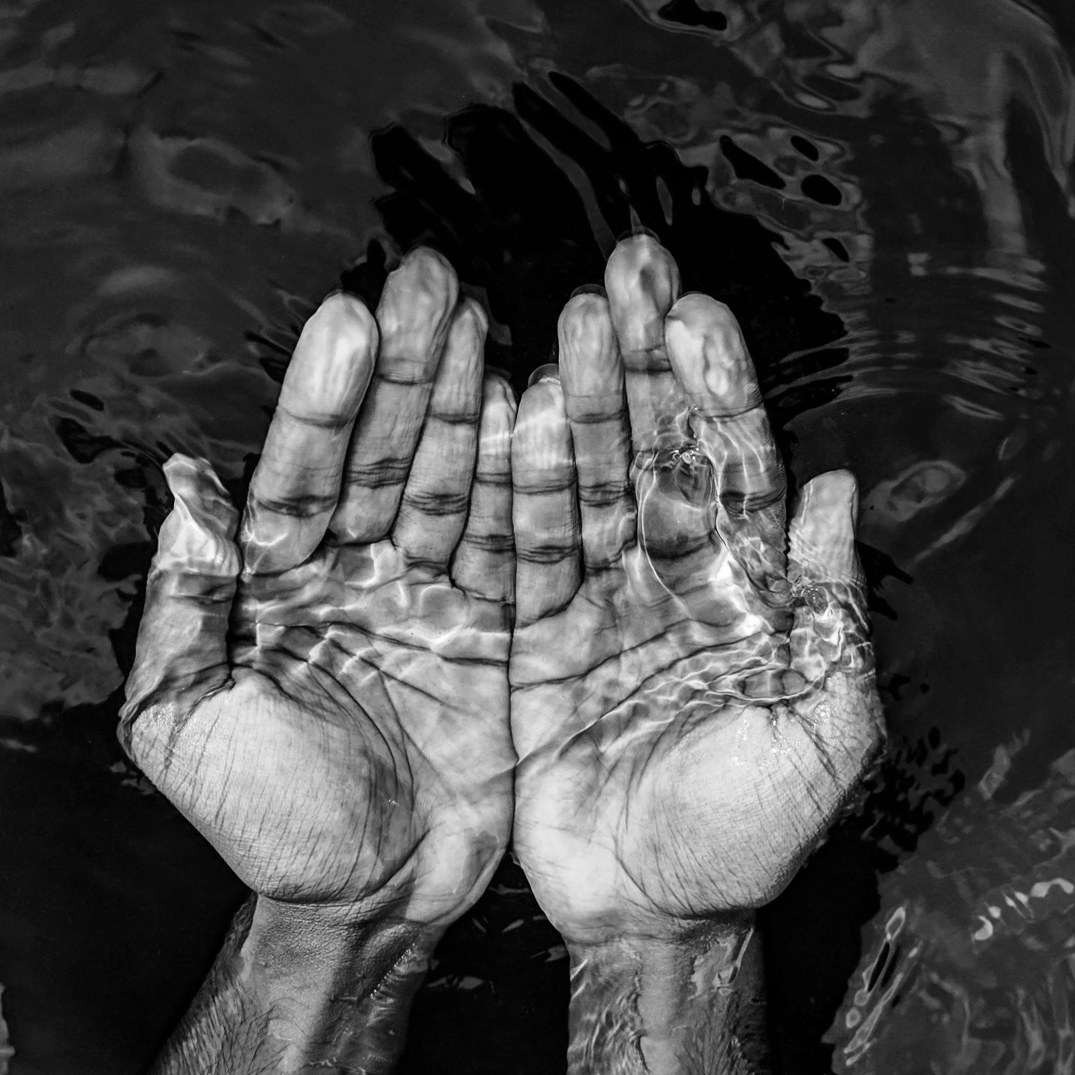 Two hands submerged in water, palms pressed together, forming a prayer-like gesture, with ripples on the water surface.