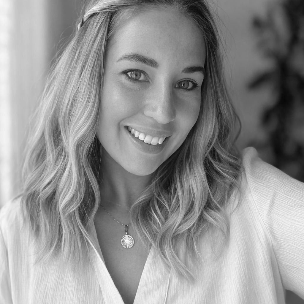 A black and white close-up photo of a smiling woman with wavy hair wearing a necklace and a light-colored top.