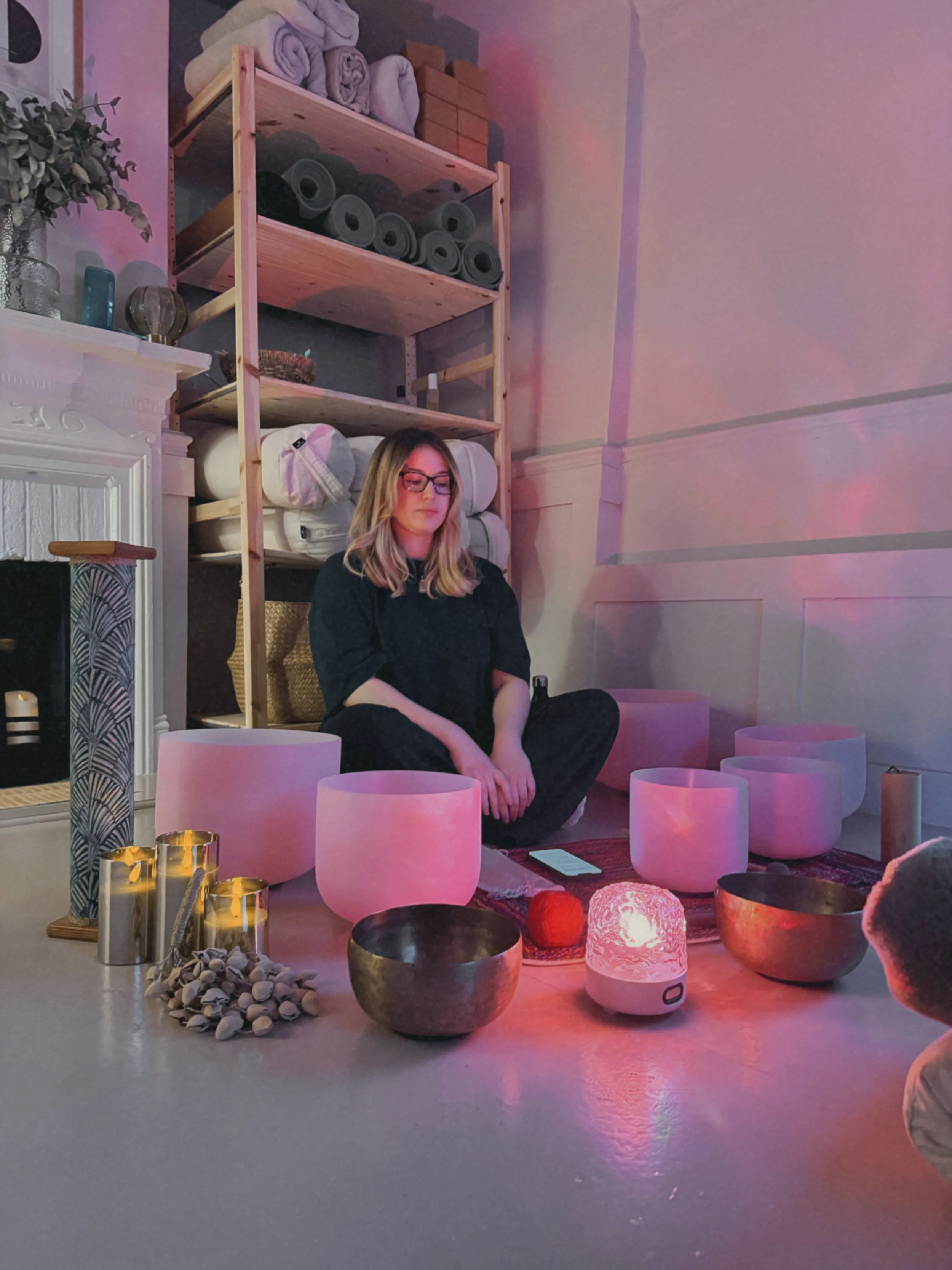 A woman sitting cross-legged on the floor in a meditation or sound healing space, surrounded by singing bowls, candles, and glowing lamps with pink and purple lighting.