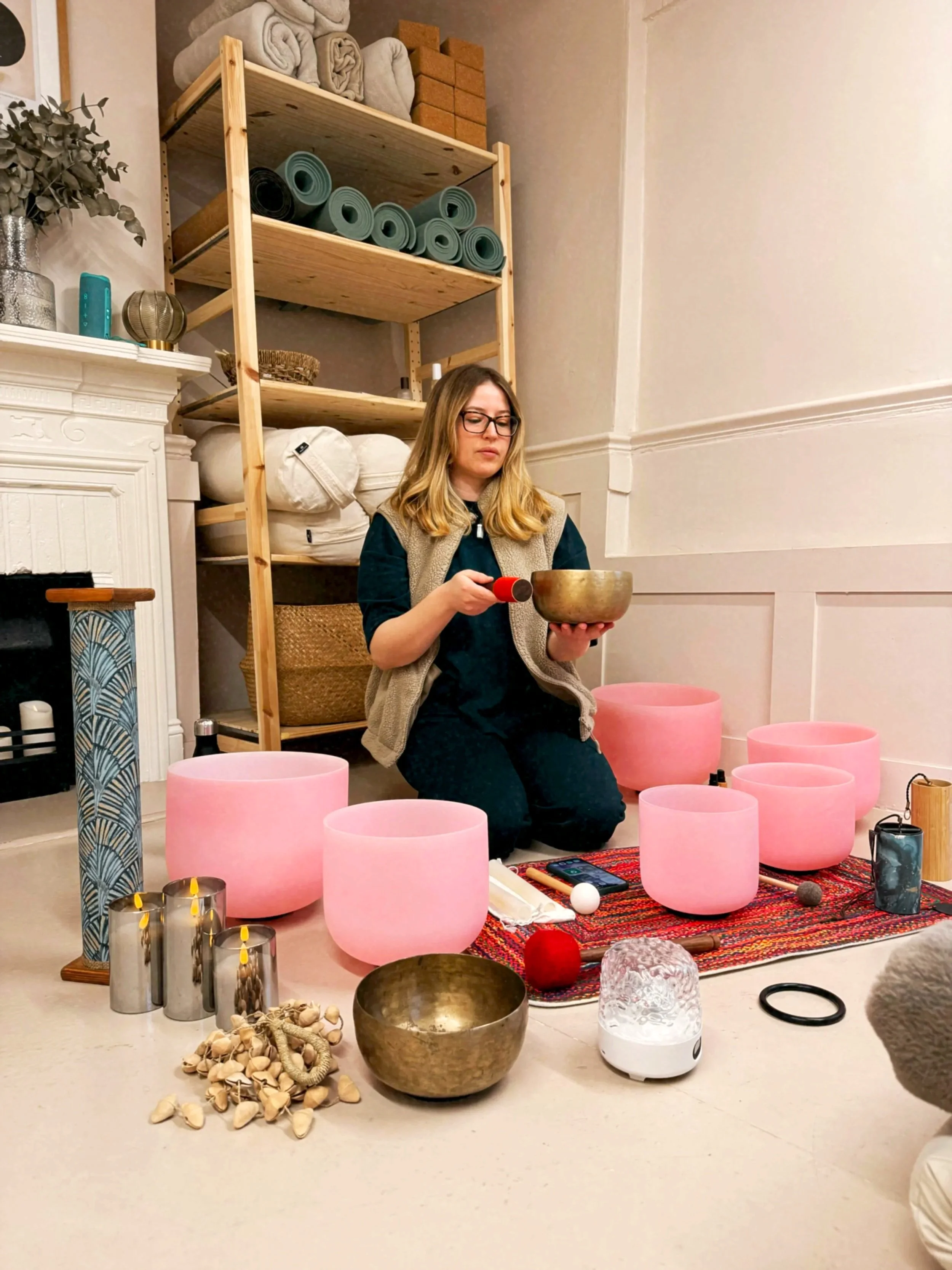 A woman sitting cross-legged on the floor, playing singing bowls with a mallet. Surrounding her are various singing bowls, candles, a small decorative object, and a colorful rug. Shelves with yoga mats, rolled towels, and storage baskets are in the background.