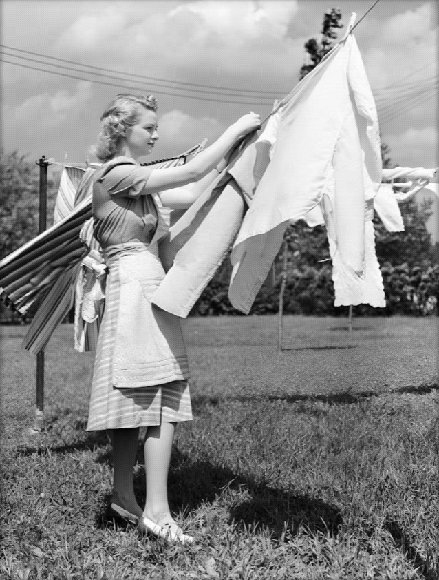 A woman hanging laundry on an outdoor clothesline in a grassy yard with trees and power lines in the background.