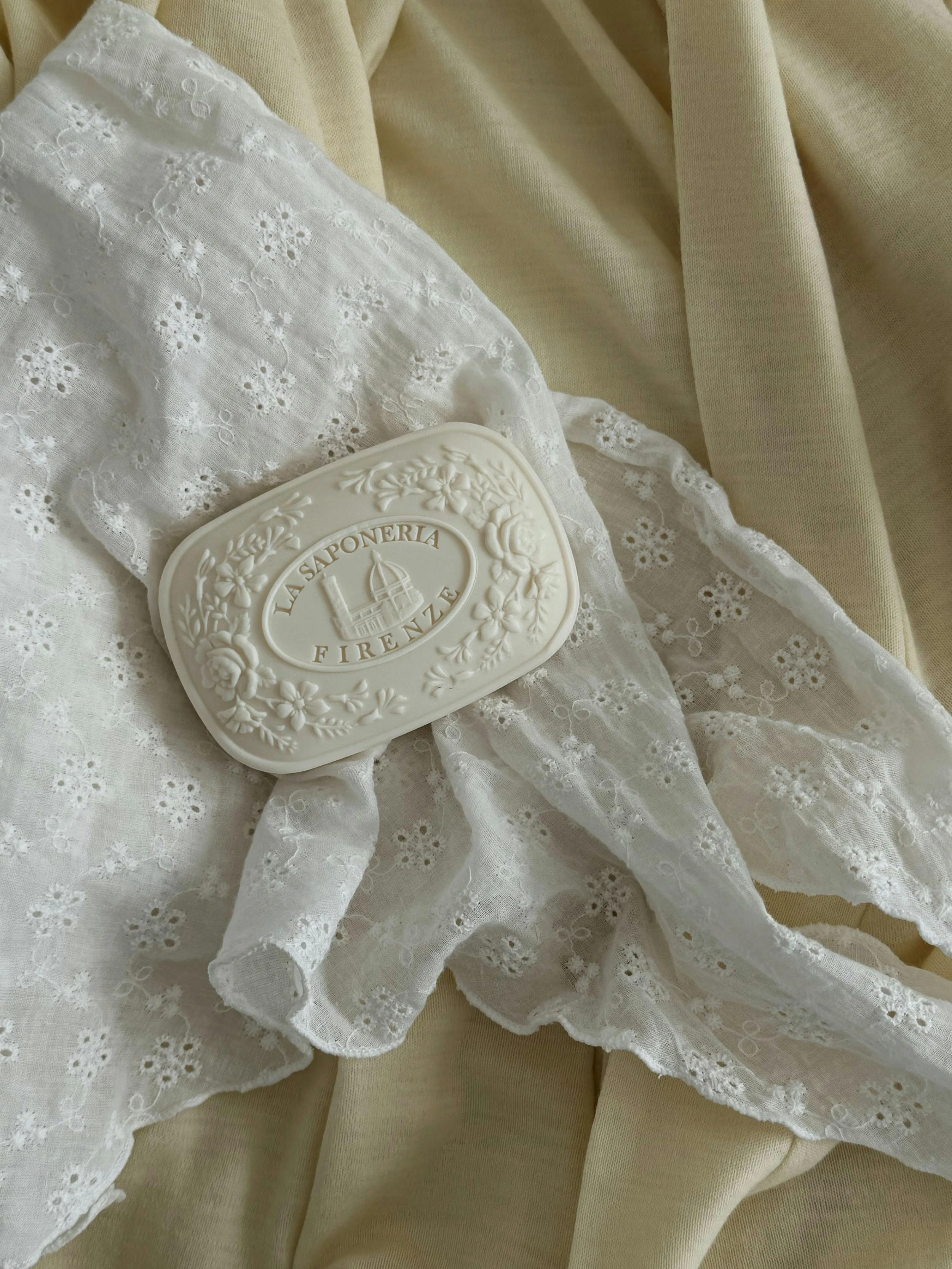 A white soap with floral embossing and the words 'La Saponeria Firenze' resting on white embroidered lace fabric, with beige fabric in the background.