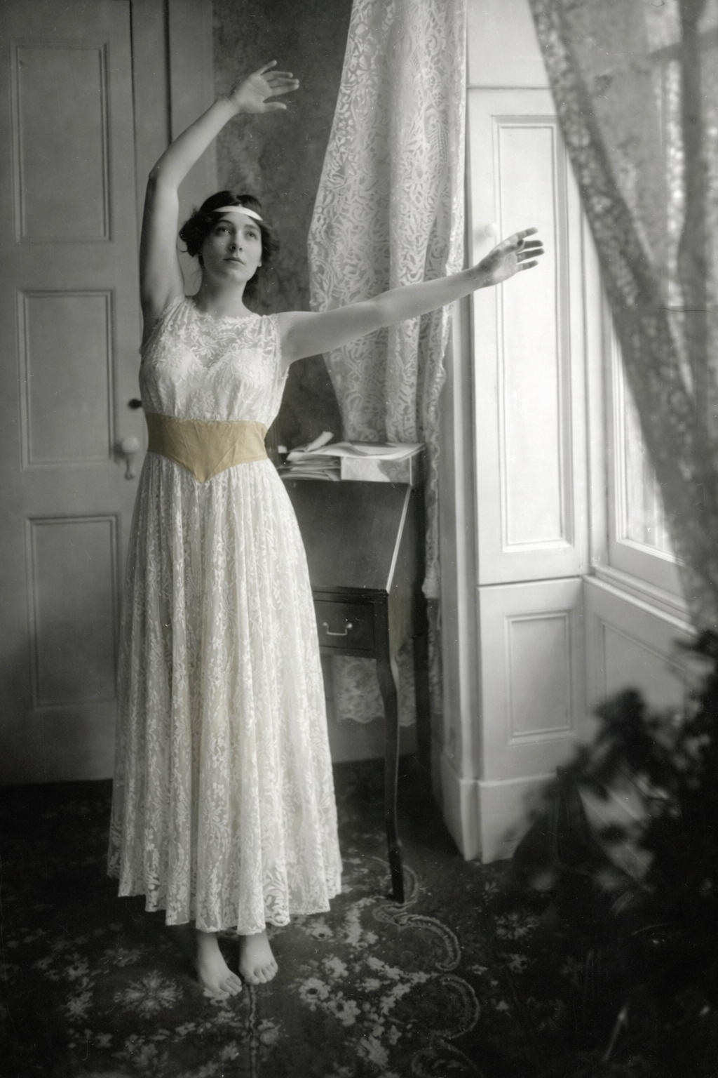 Black and white photo of a woman in a long, light-colored lace dress, standing indoors near a window with patterned curtains, with her arms raised in a ballet pose.