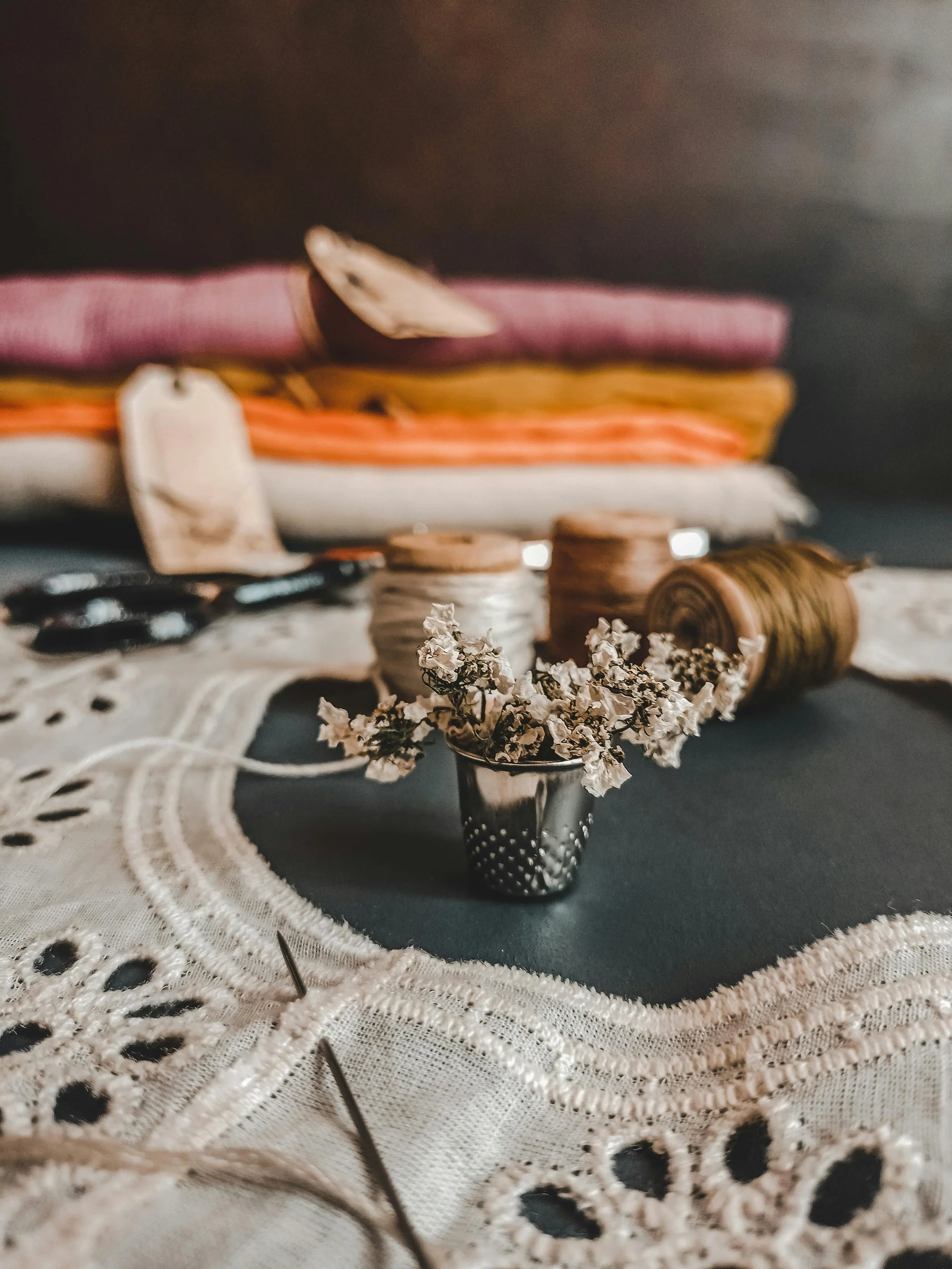 A small silver vase with white dried flowers on a white embroidered cloth, with spools of thread, scissors, and folded fabric in the background.