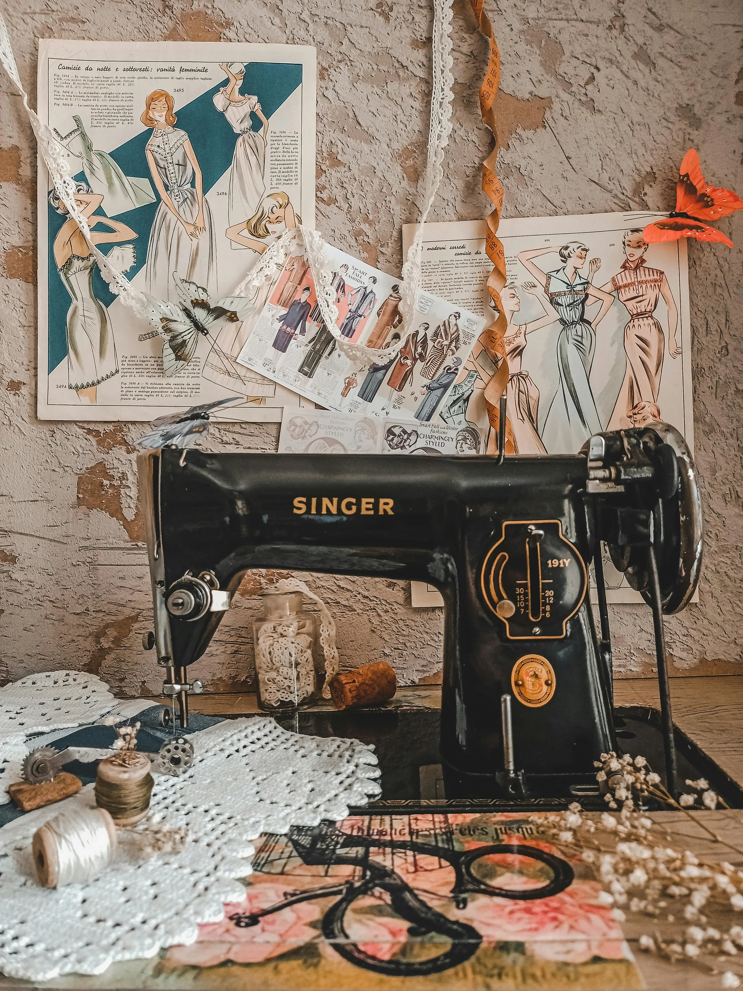 Vintage Singer sewing machine on a cluttered table with fabric, thread, and small decorative items, set against a textured wall with fashion illustrations and sewing patterns pinned up.