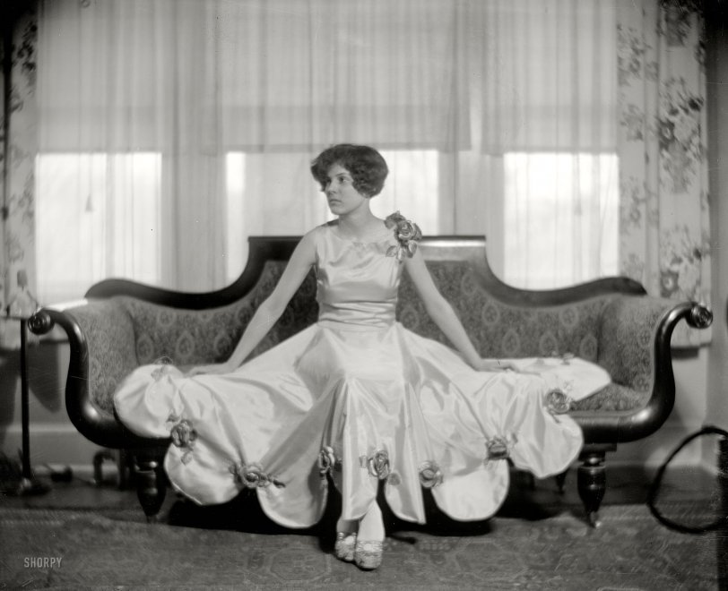 A woman in an elegant dress sits on a vintage sofa in a room with floral curtains and a carpet.