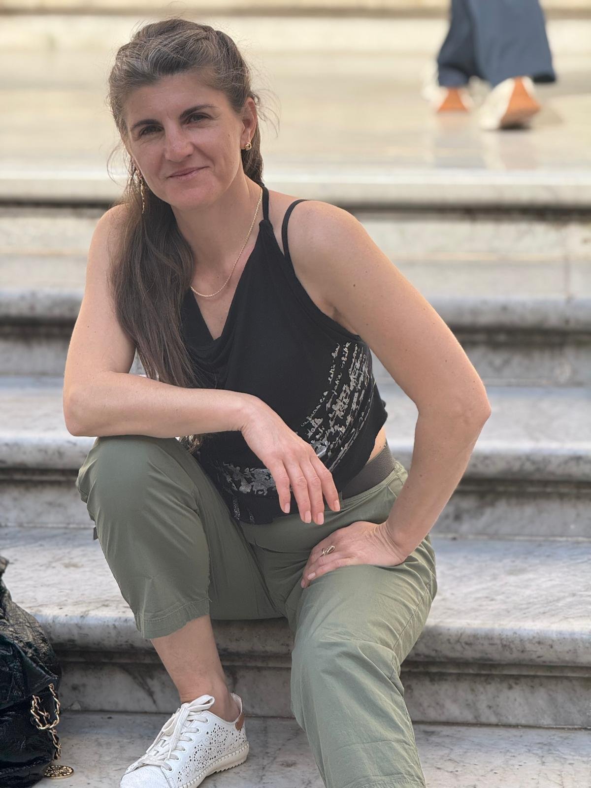 Una donna seduta su delle scale di marmo, indossa un top nero, pantaloni kaki e scarpe bianche. Tiene una mano sulla gamba e sorride leggermente.