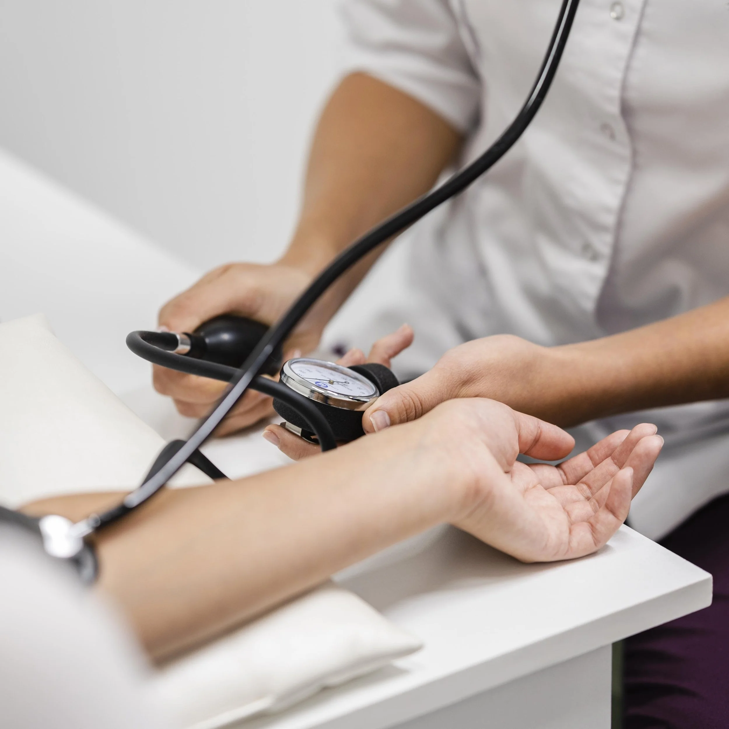 Un medico o un infermiere controlla la pressione arteriosa di un paziente utilizzando uno sfigmomanometro, mentre il paziente si appoggia su un tavolo bianco.