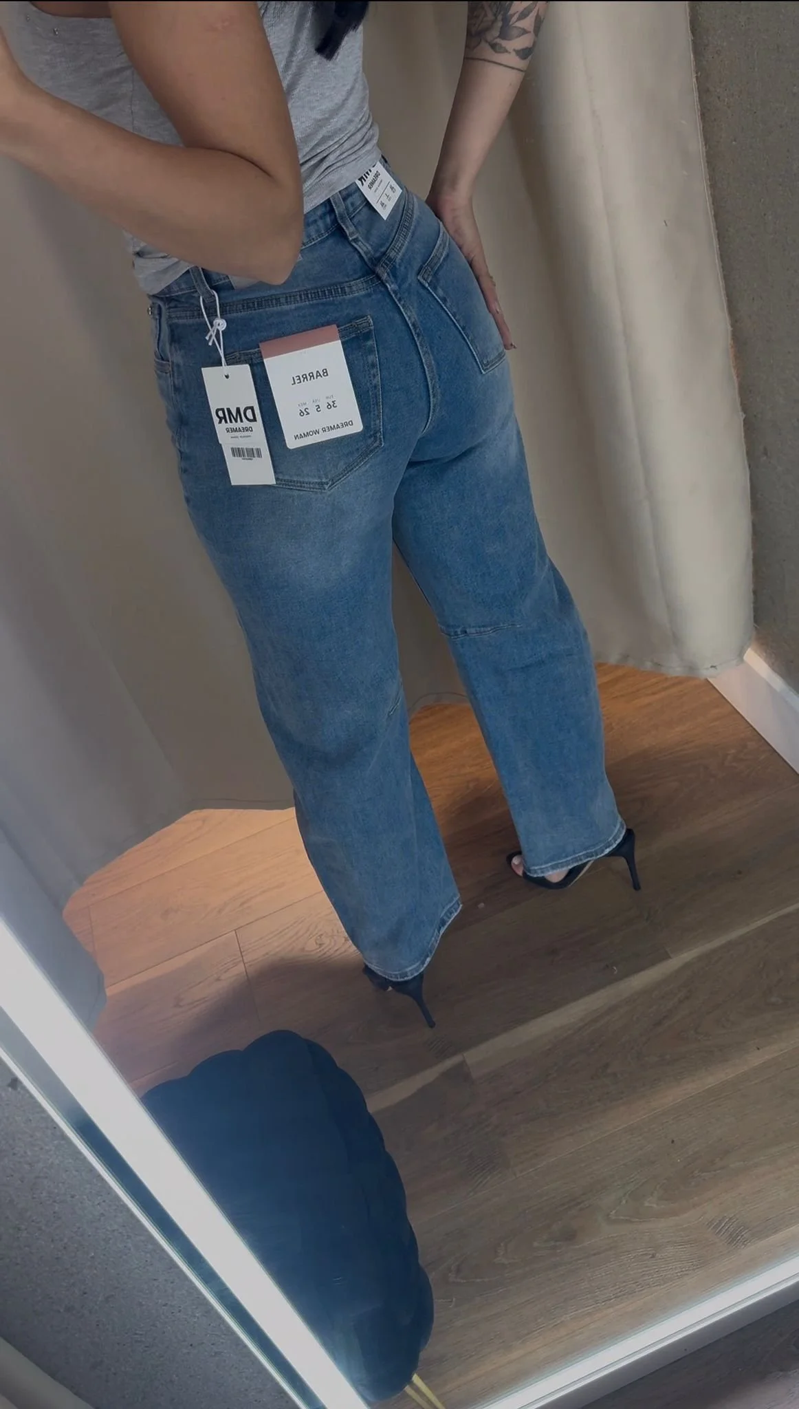 Person wearing blue jeans and black high heels, taking a mirror selfie in a fitting room, with tags visible on the jeans.
