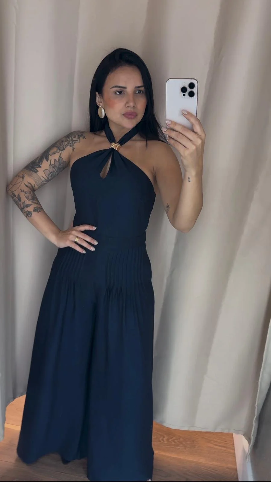 Woman taking a mirror selfie wearing a black halter dress with a keyhole neckline and wide-legged pants, standing against a beige curtain.