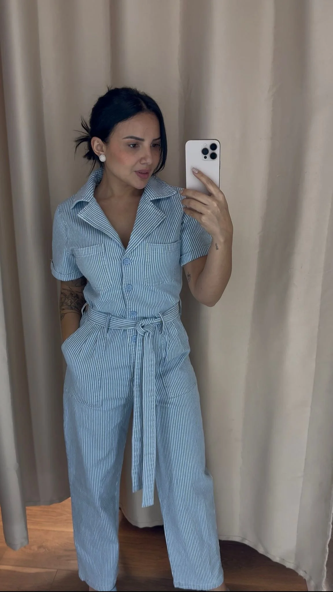 A woman taking a mirror selfie in a room with beige curtains. She is wearing a light blue and white striped short-sleeve jumpsuit with a collar and a waist tie, and has tattoos on her arms. She has dark hair pulled back and is wearing large white ear