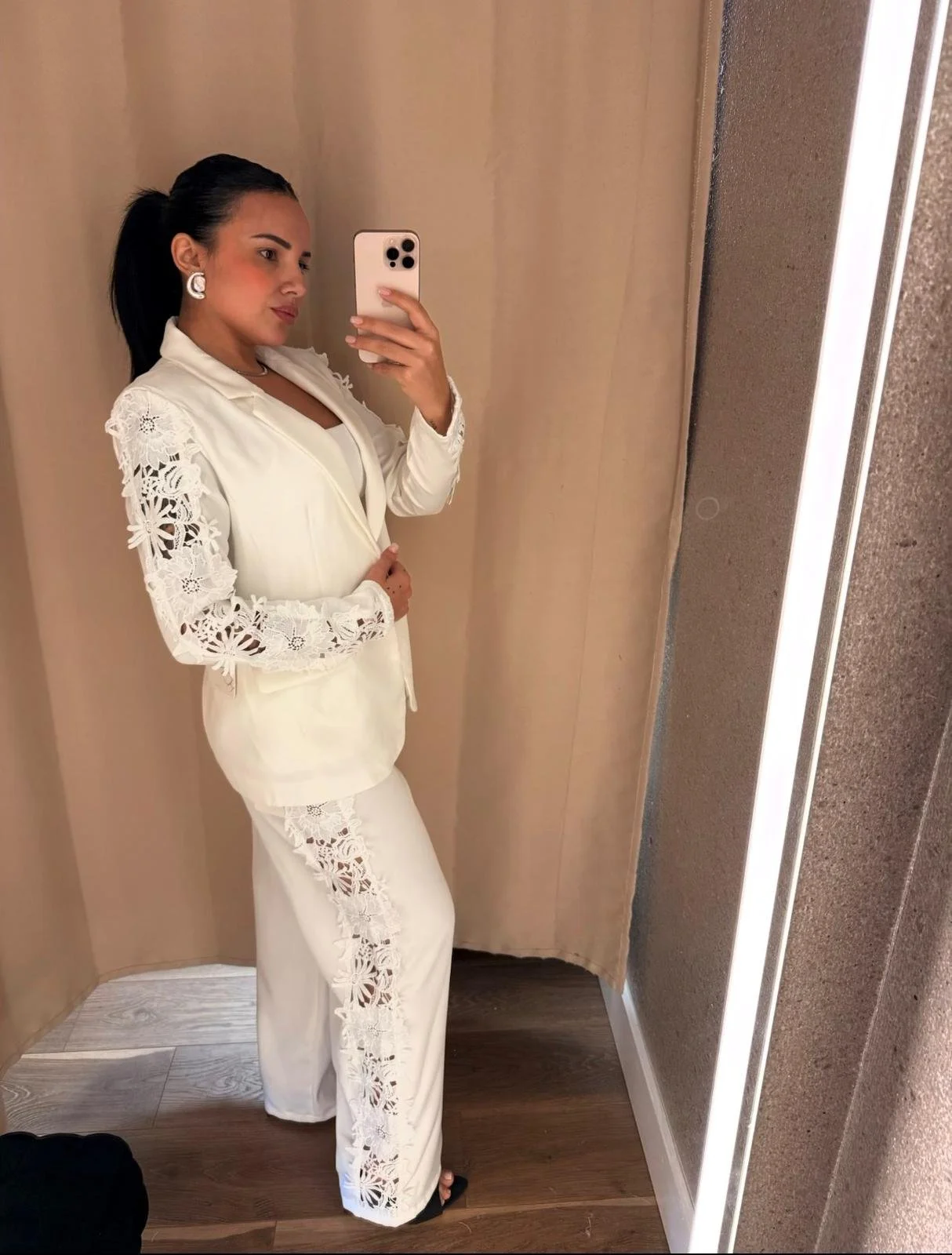 Woman taking a mirror selfie in an elevator, wearing a white outfit with lace details, with dark hair pulled back and wearing earrings.