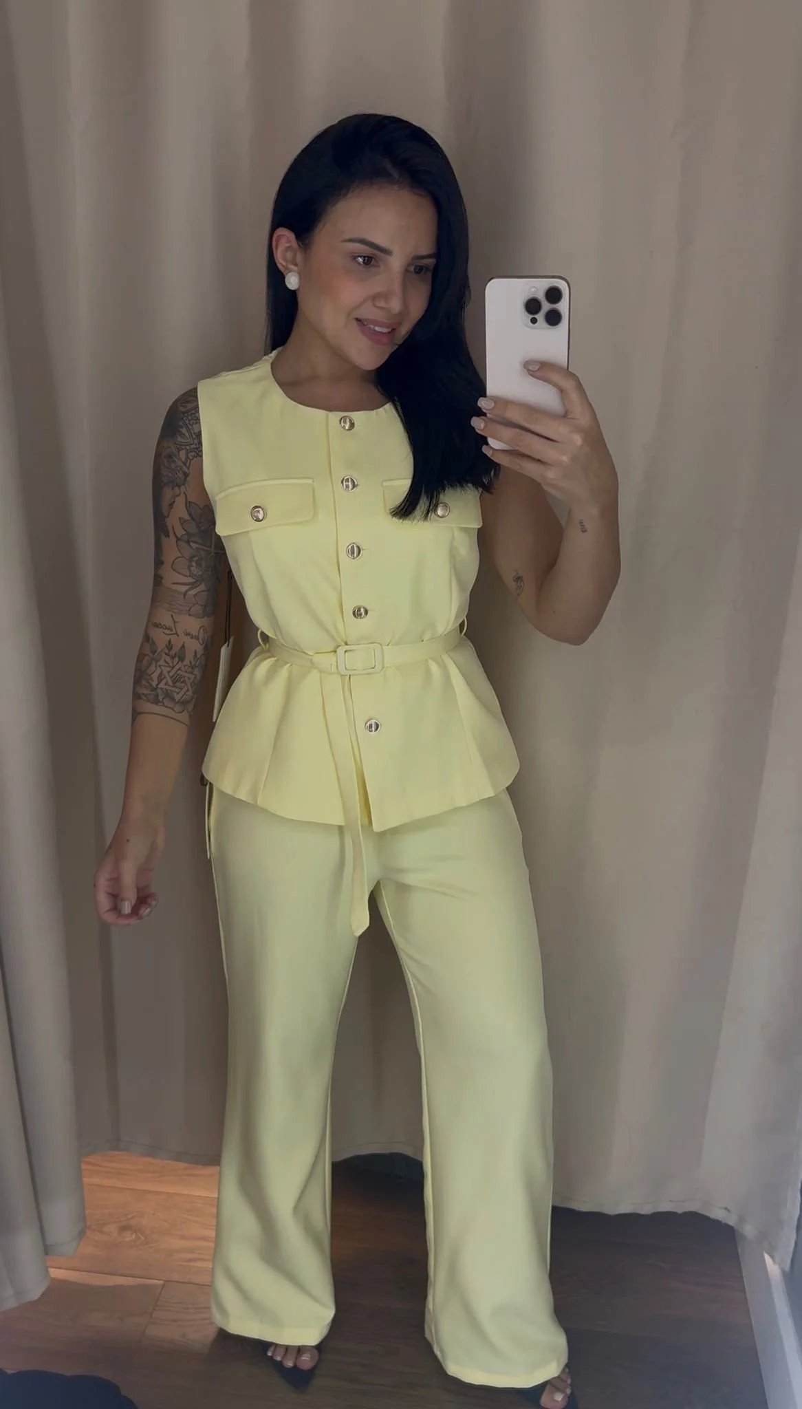 Woman in light yellow sleeveless button-up top and matching wide-leg pants, taking a mirror selfie in a fitting room.