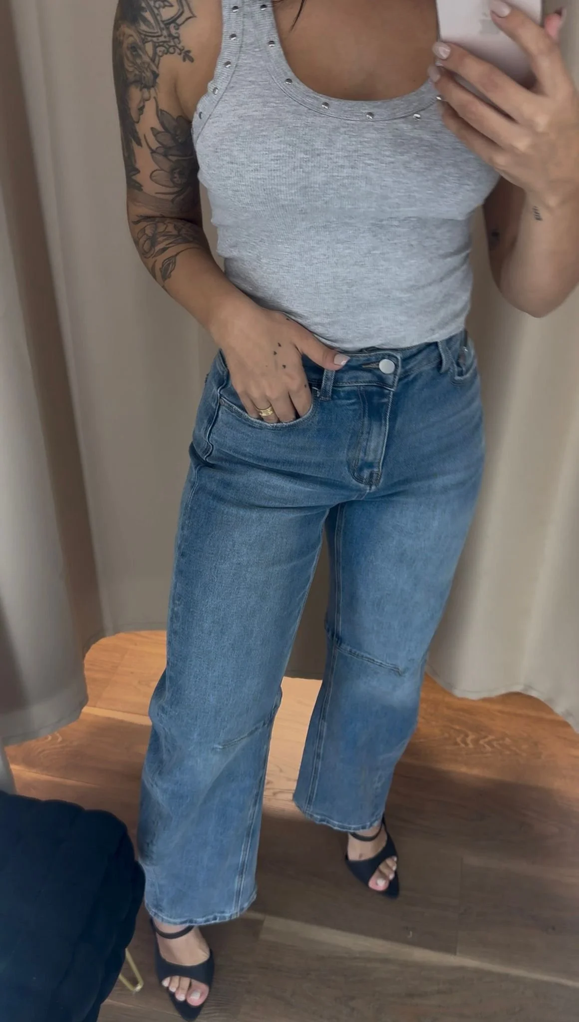 A woman taking a mirror selfie showing her gray tank top with studded details, blue jeans, and black high-heeled sandals. She has tattoos on her left arm and hand.