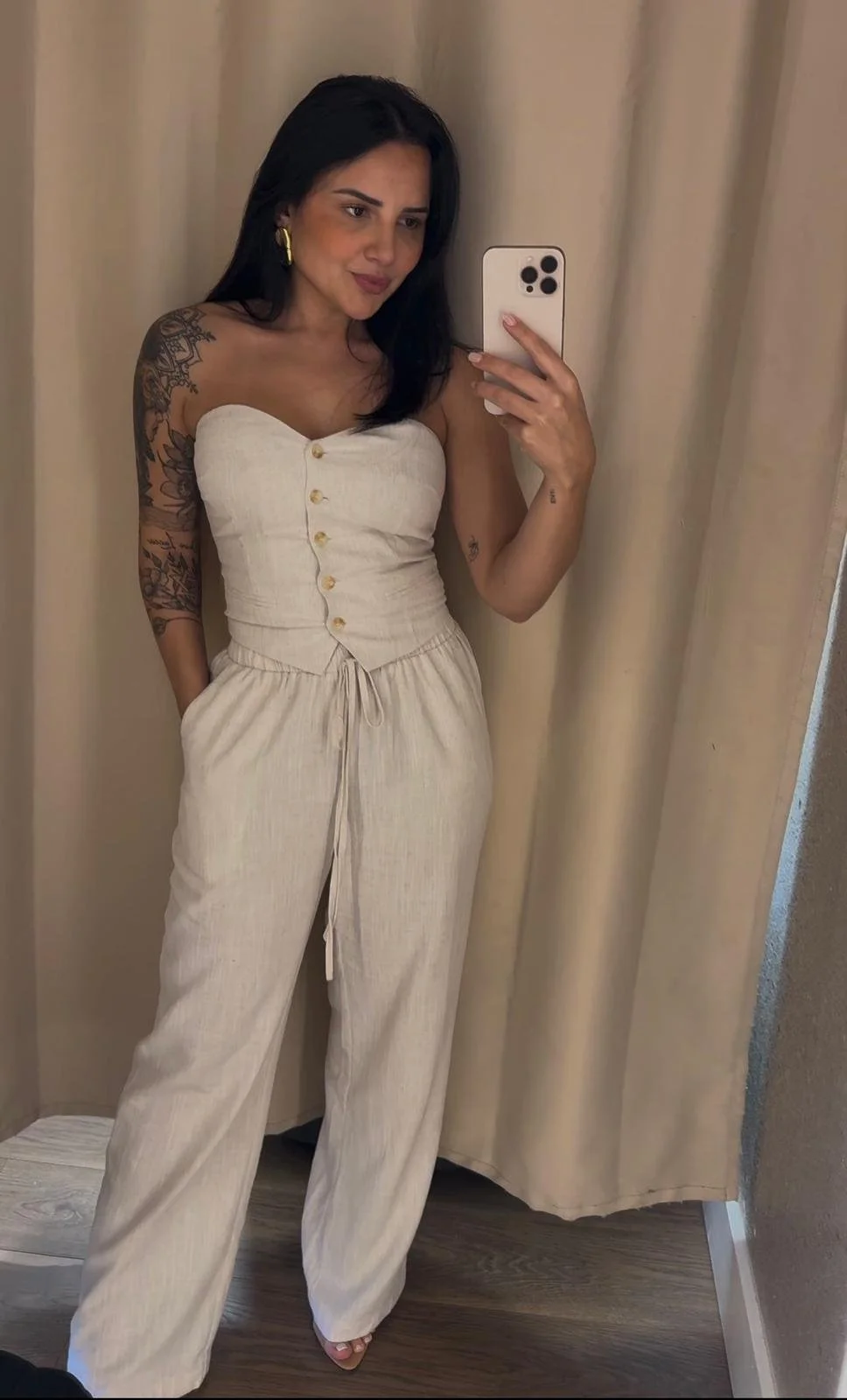 A woman with dark hair and tattoos wearing a beige strapless jumpsuit taking a mirror selfie.