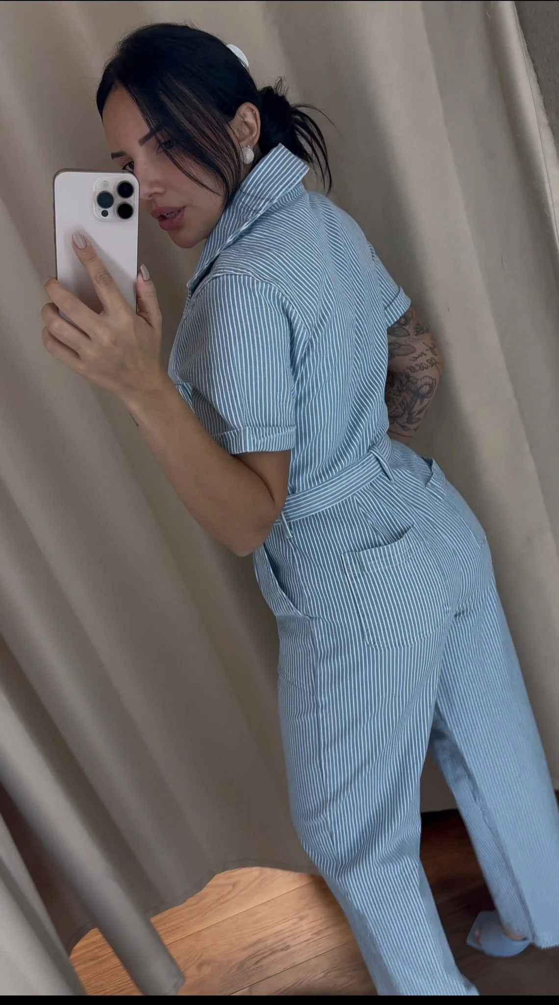 Woman taking a mirror selfie in a light blue and white pinstripe jumpsuit, showing her face and tattoo on her left arm.
