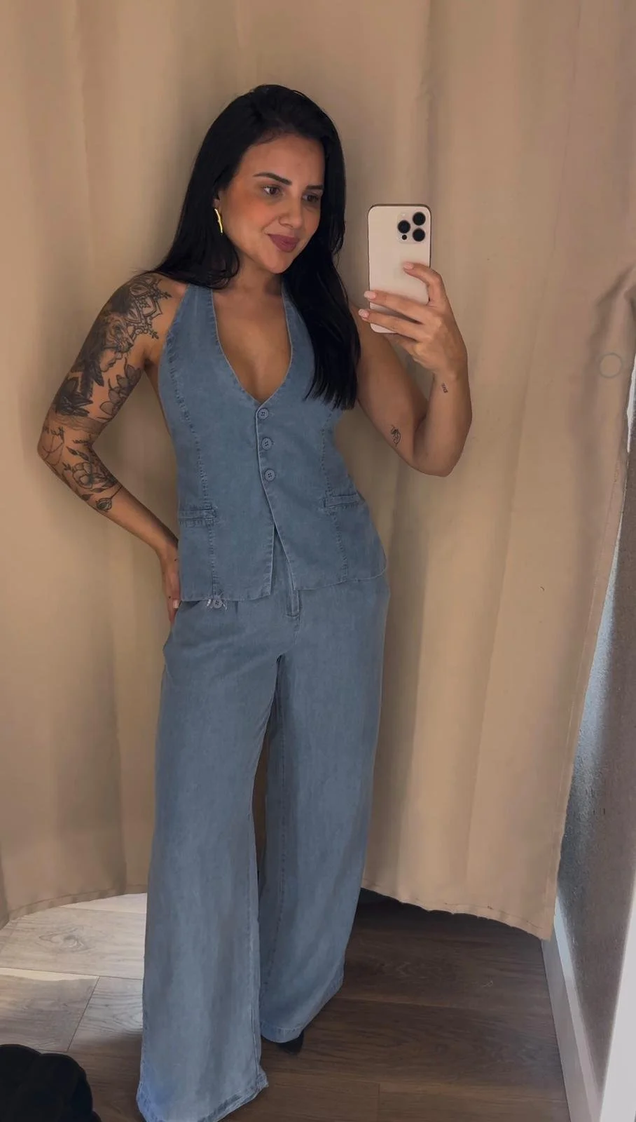 A woman taking a mirror selfie with a smartphone, wearing a denim sleeveless button-up top and matching wide-leg jeans. She has dark hair, gold hoop earrings, and visible tattoos on her left arm.