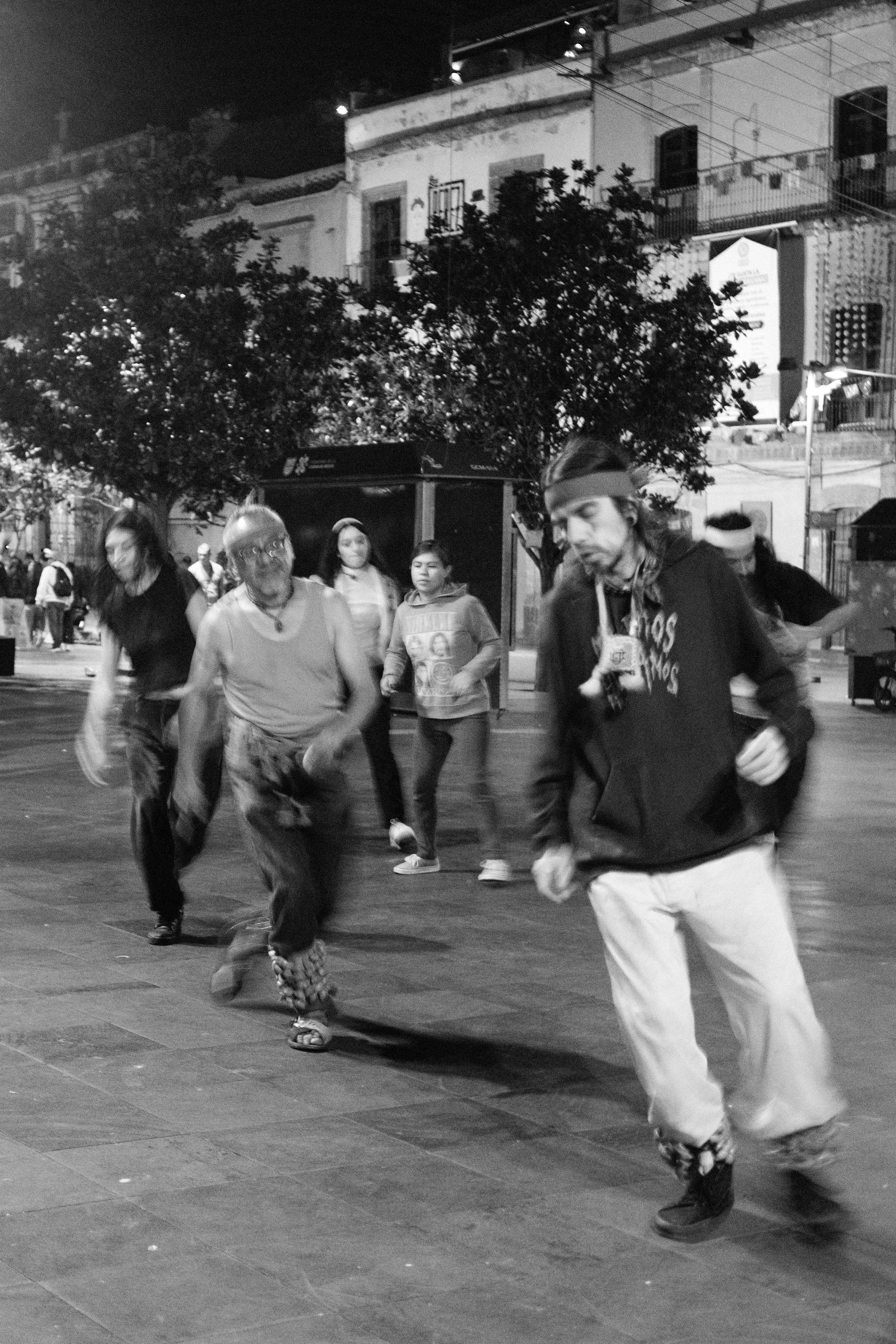 People dancing and socializing in an outdoor urban space at night, with buildings and trees in the background.