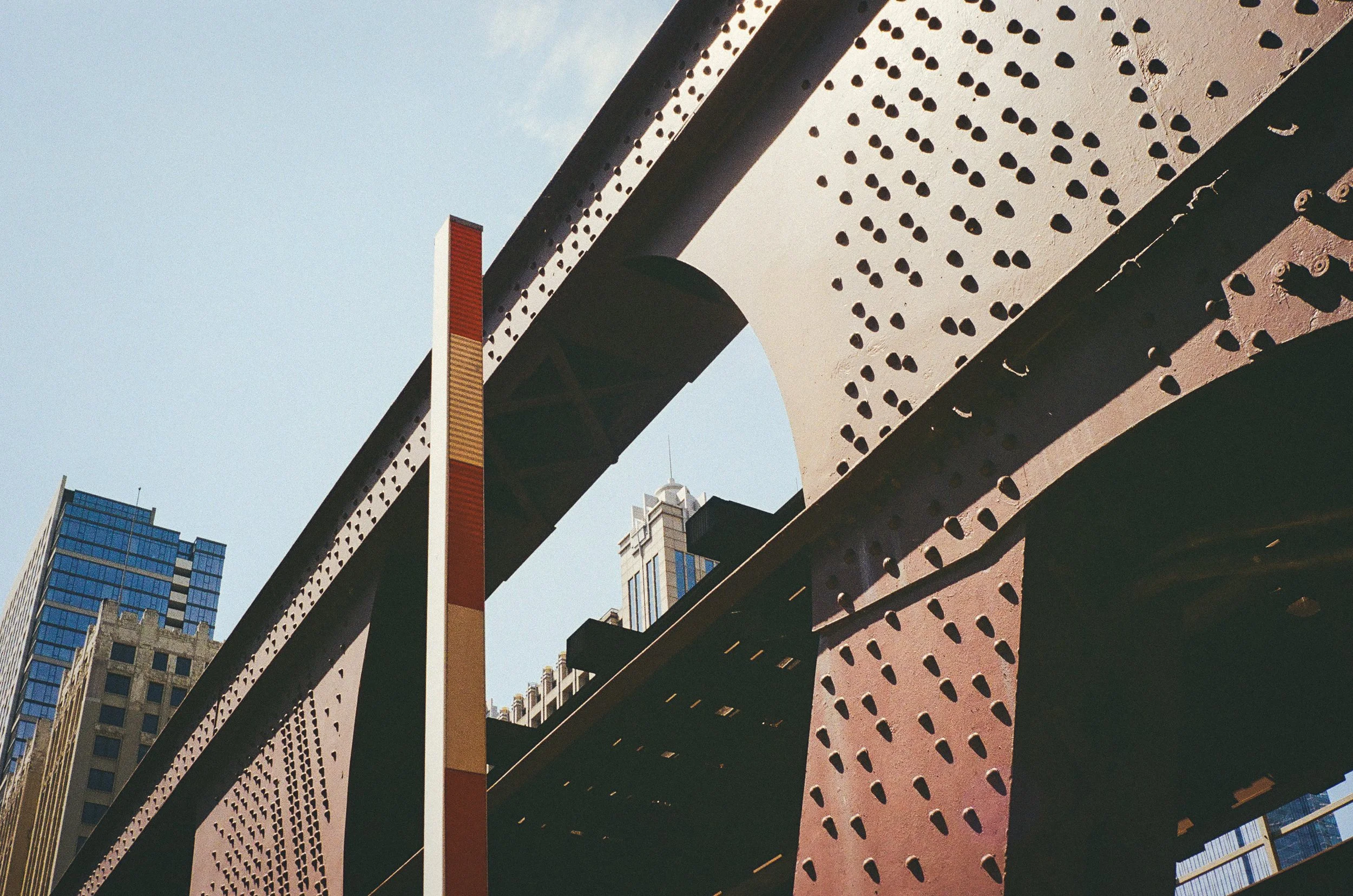 Underside of a large steel bridge with rivets, high-rise buildings in the background, and a clear blue sky.