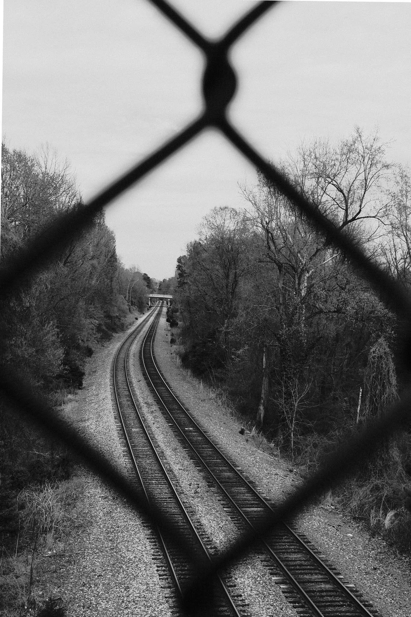 Black and white photograph of railway tracks viewed through a chain link fence with trees on either side.