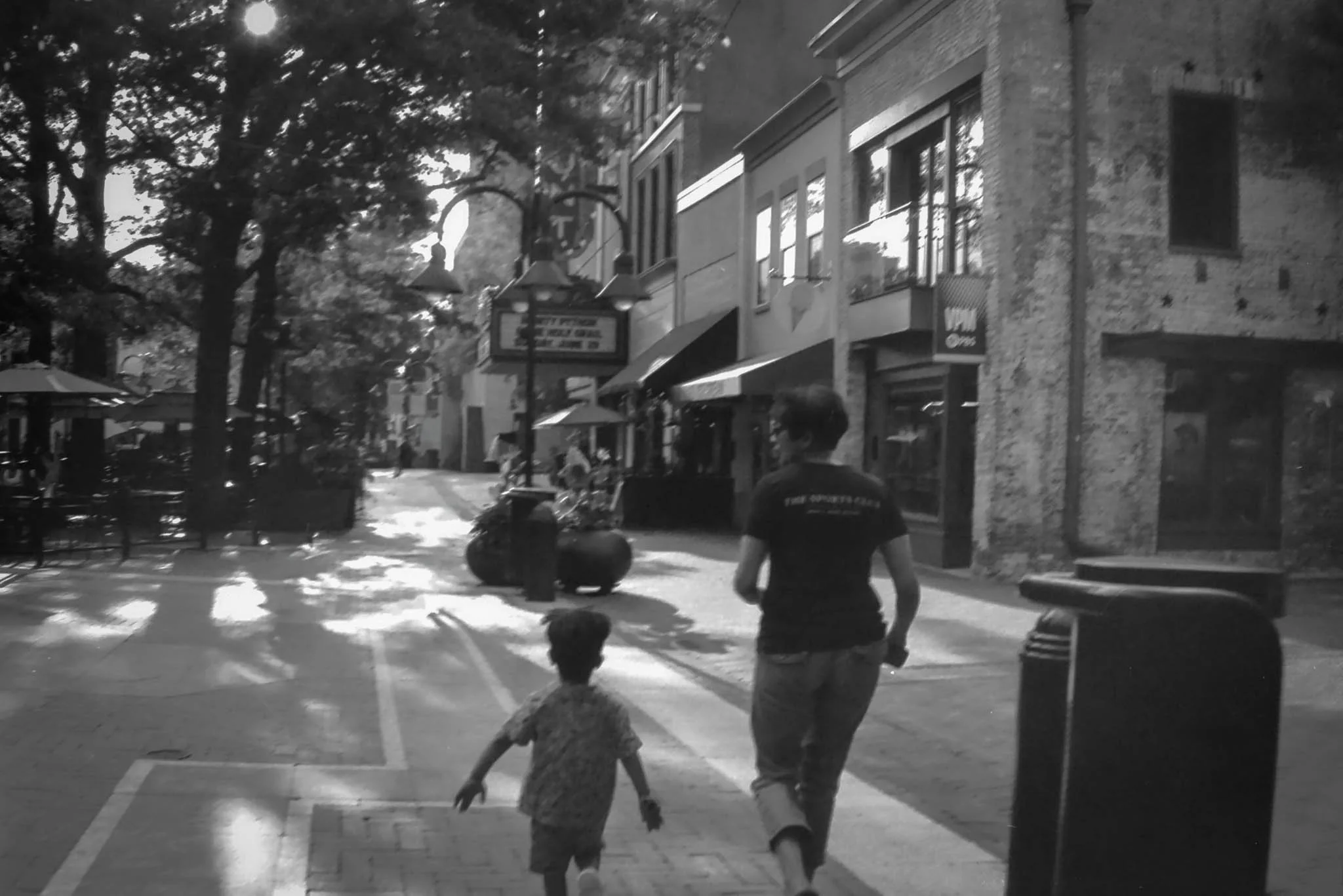 A woman and a young boy walk hand in hand along a city sidewalk lined with trees, shops, and outdoor seating areas, with sunlight filtering through the trees.