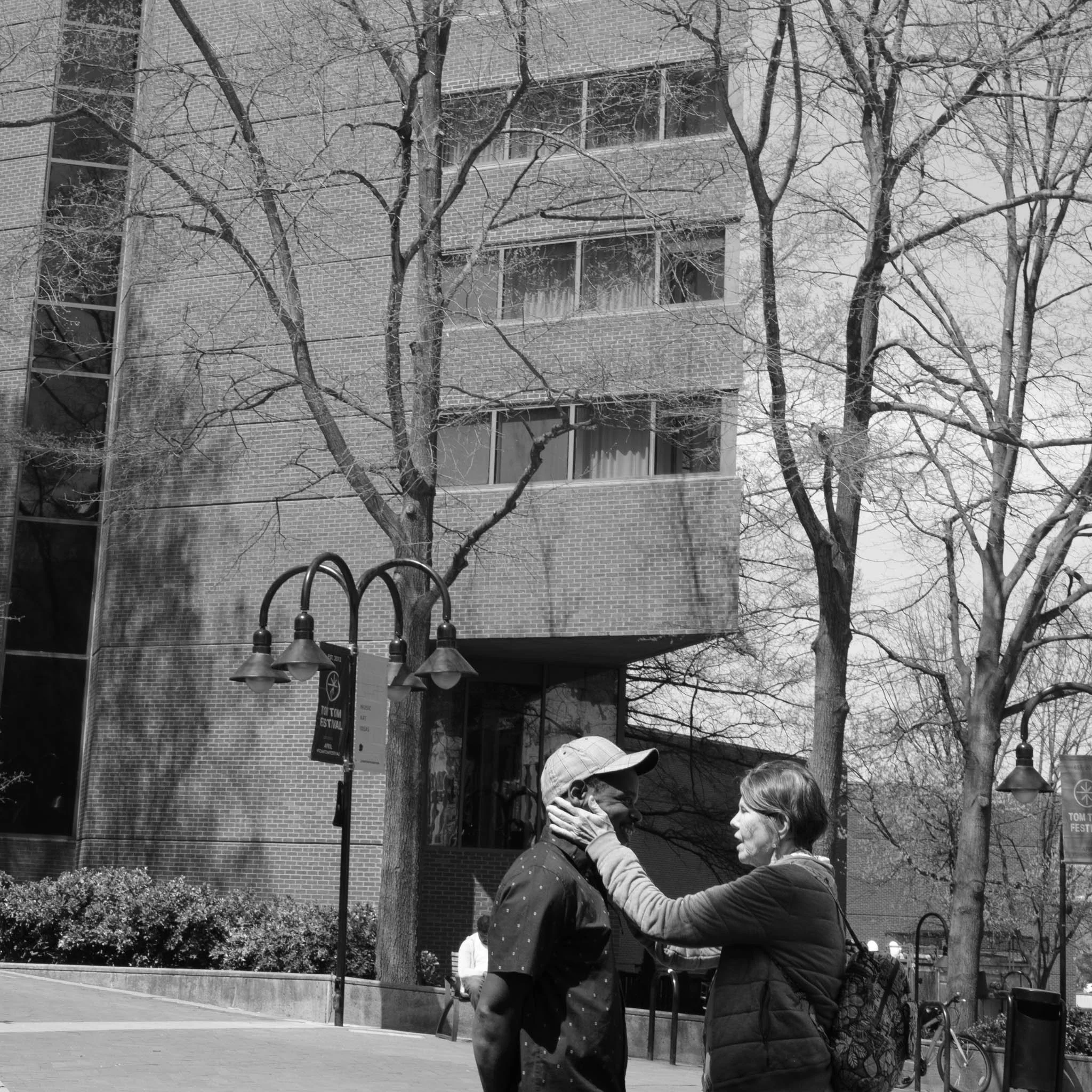 A woman and a man facing each other outdoors, with the woman touching the man's face, in front of a brick building and leafless trees.