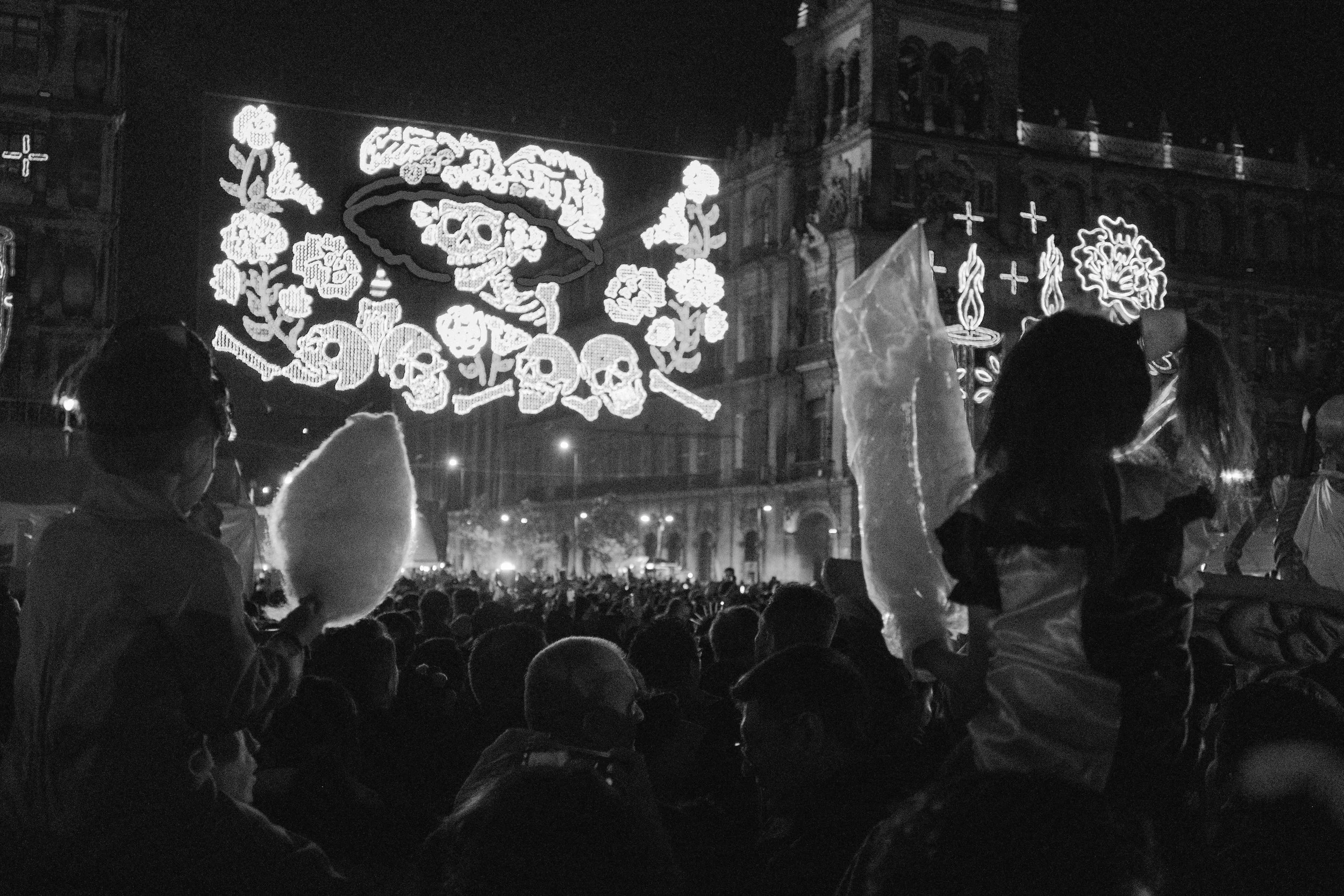 Crowd of people at a nighttime event with illuminated decorations hanging above, including skulls, flowers, and candles, and a large historic building in the background.