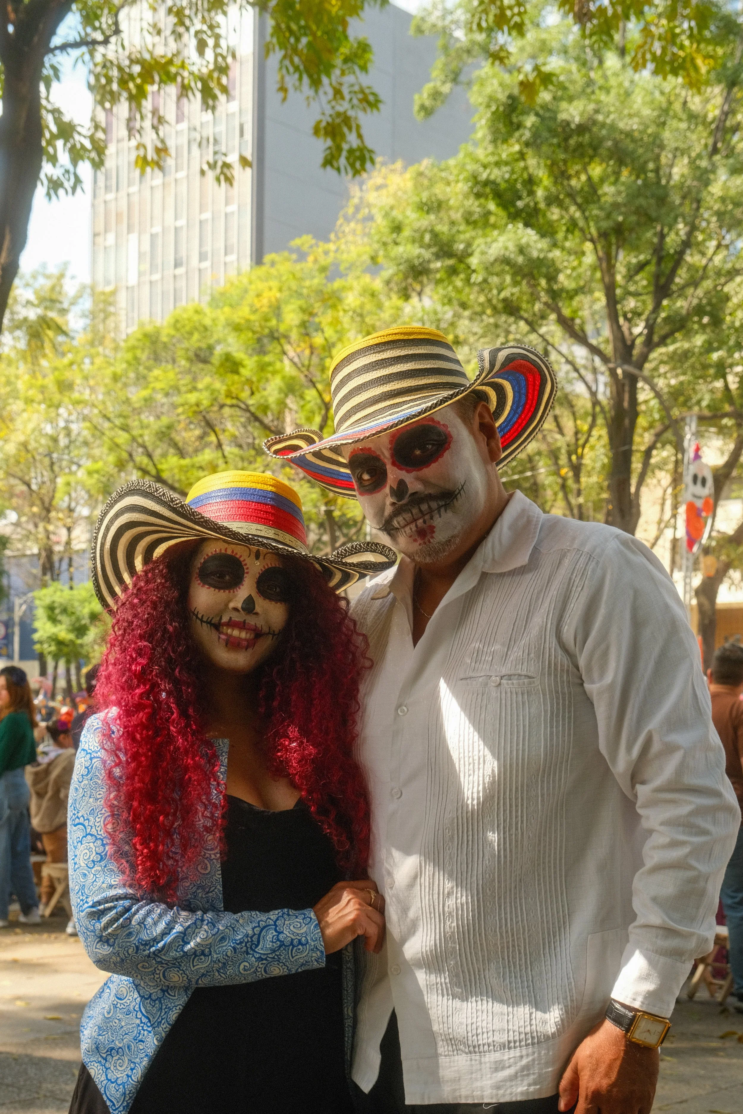 Two people dressed in traditional Mexican sugar skull costumes with painted faces, wearing striped sombreros, standing outdoors among trees and other people during a celebration.