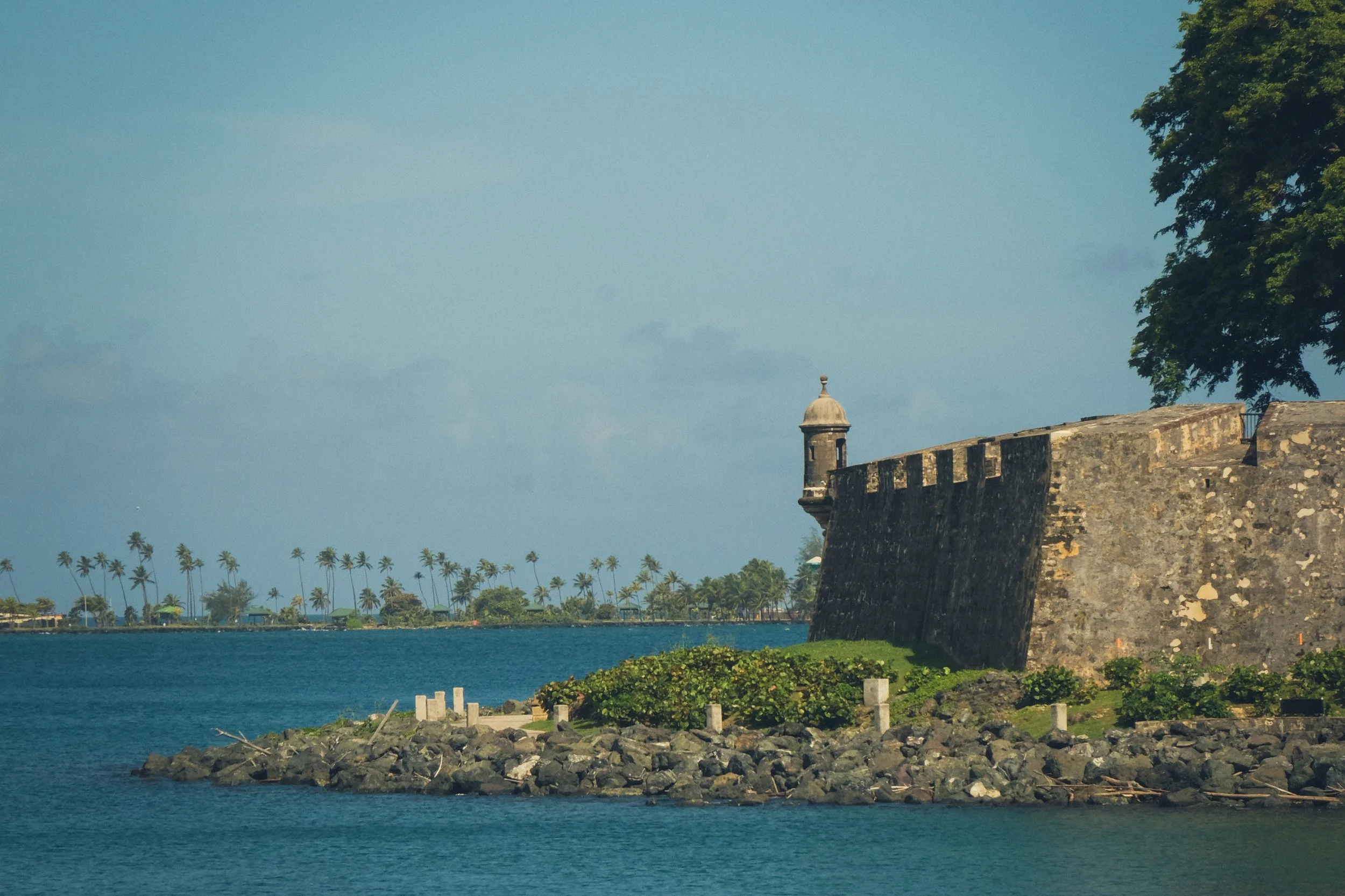 Fortified stone wall by the water with a small turret and lush greenery, set against a tropical backdrop with palm trees and a cloudy sky.