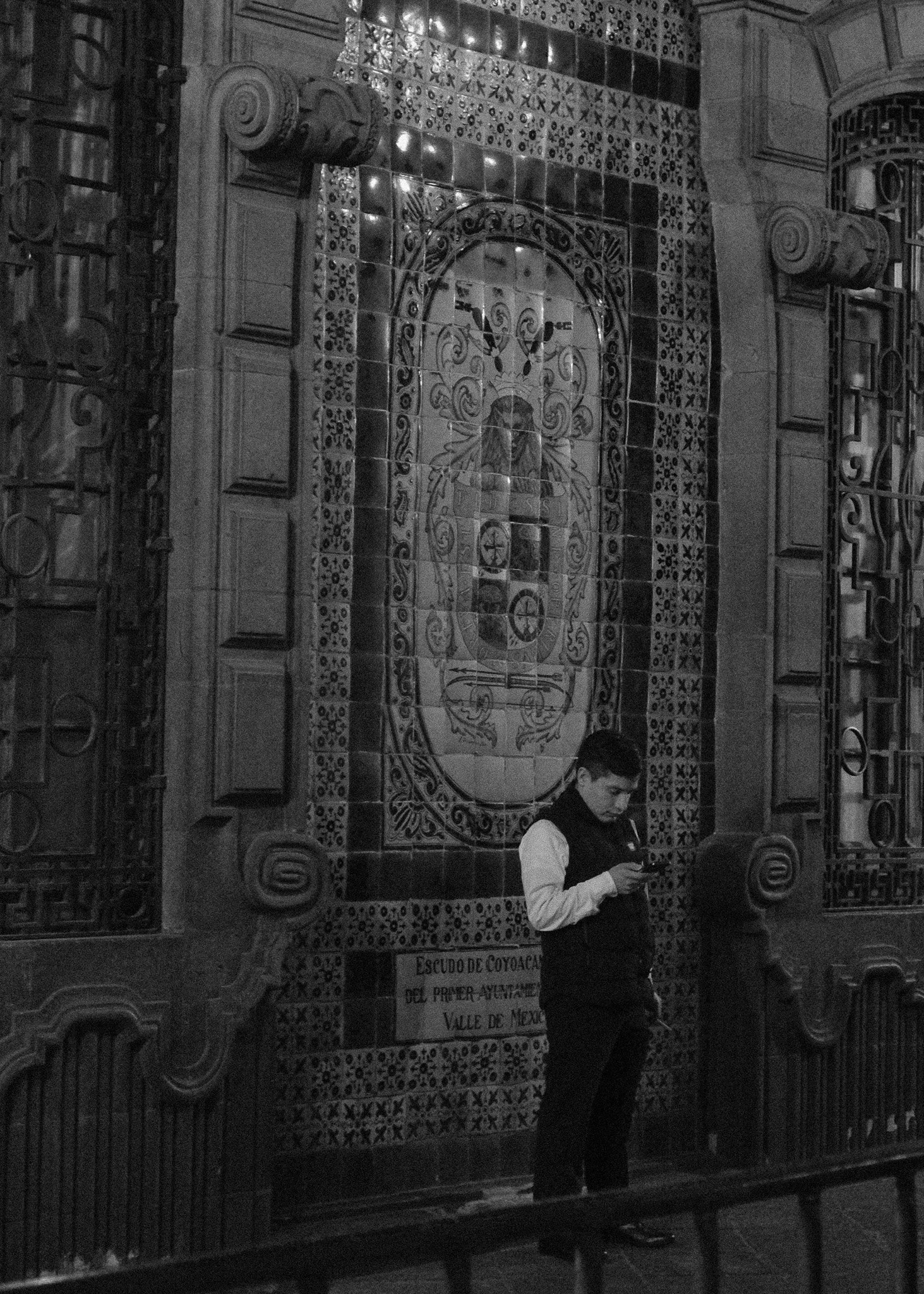 A young man standing and looking at his phone in front of a decorated tile mural with religious symbols and text in Spanish, in what appears to be a church or religious building.