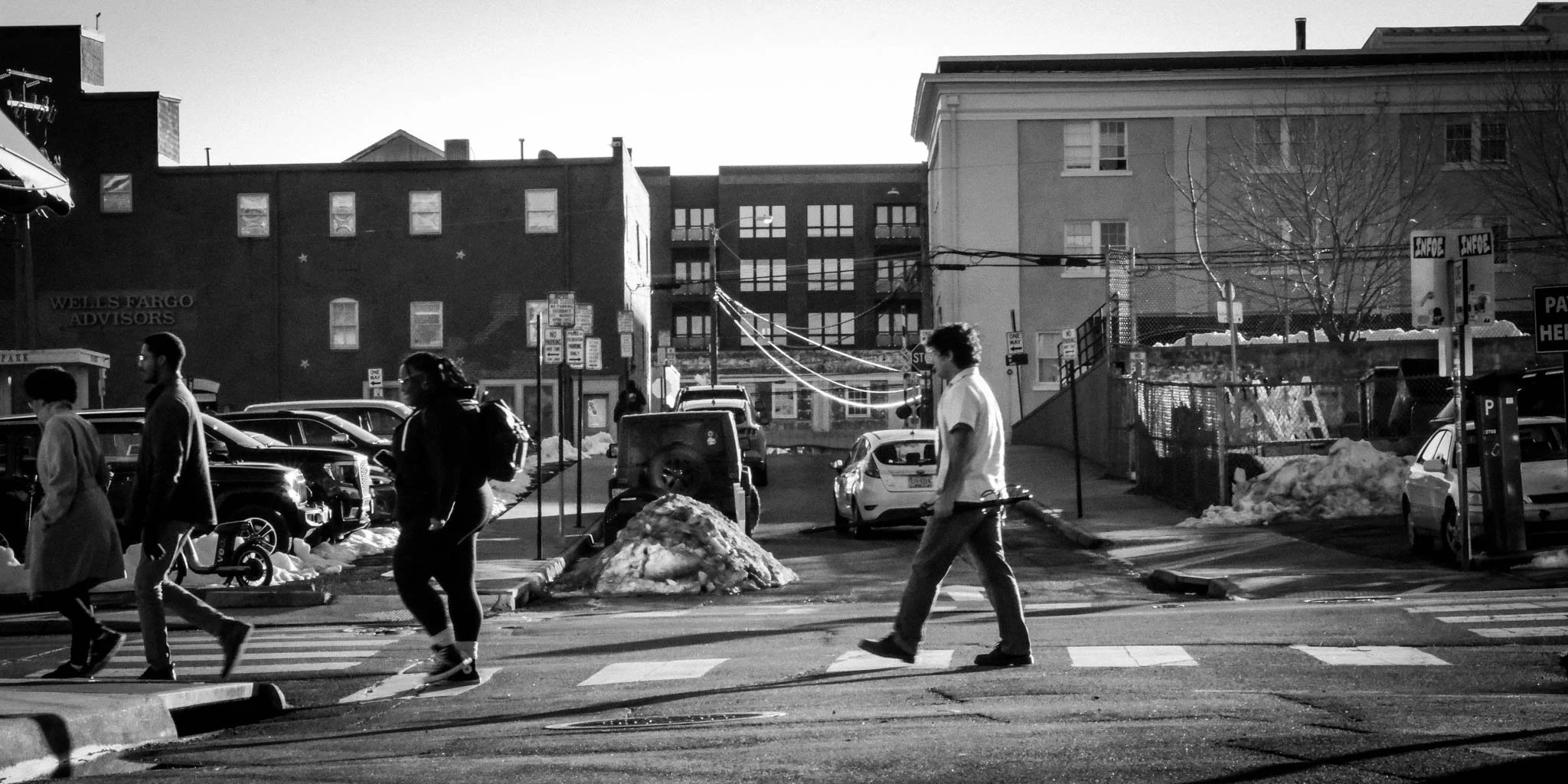 A black and white photo of people crossing a city street with parked cars, snow piles on the sides, and buildings in the background.