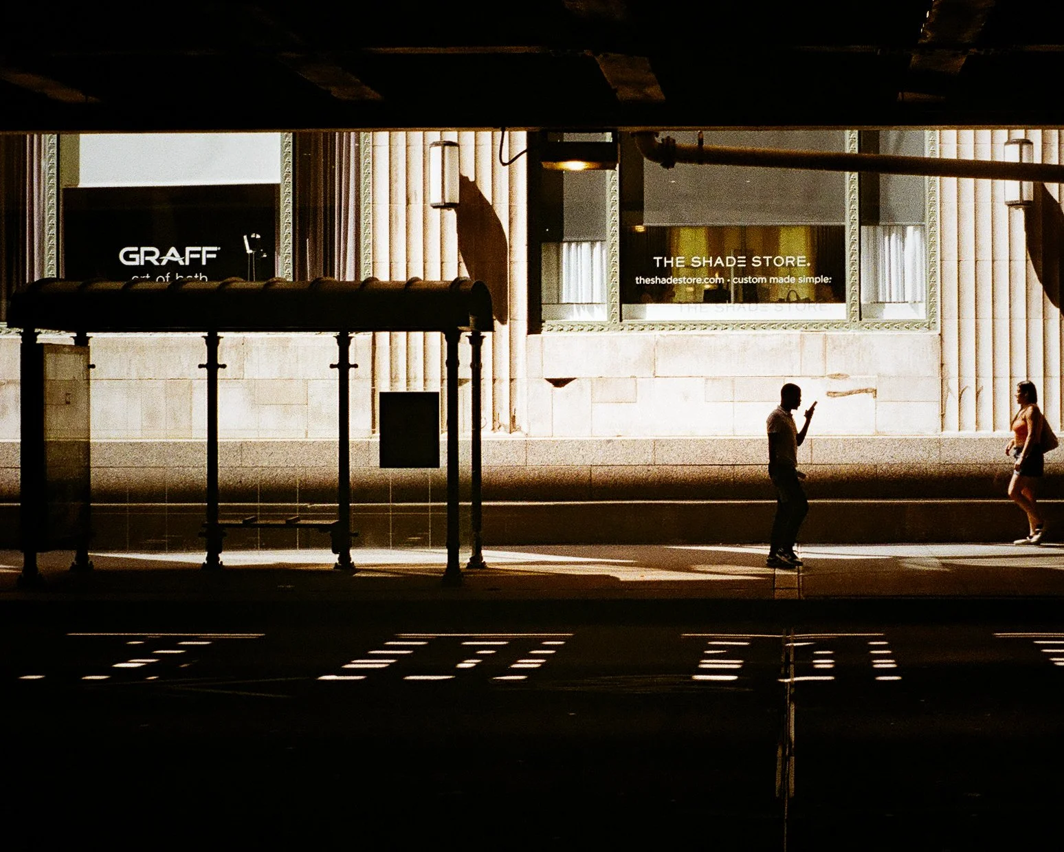 Silhouetted man and woman walking on city sidewalk under an overpass, illuminated by sunlight, with storefronts and advertisements in the background.