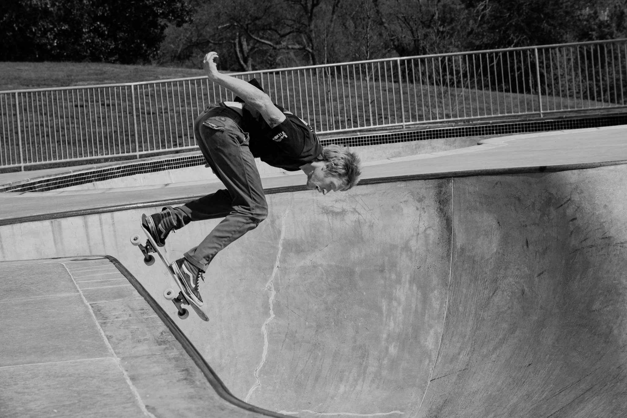 A young man skateboarding in a concrete skatepark bowl, captured mid-ride with an expression of excitement, black and white photograph.