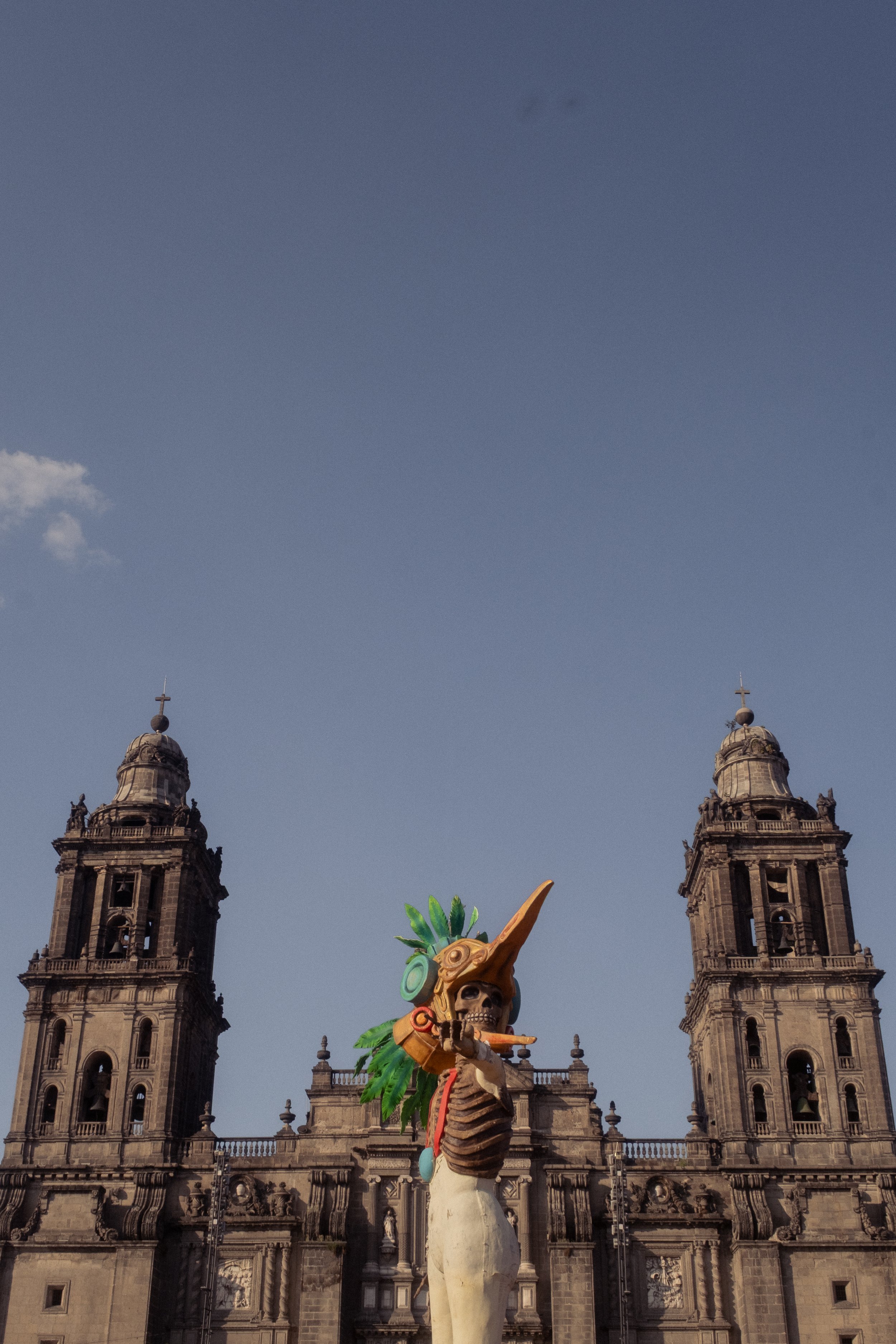 A colorful sculpture of a skeleton with a large orange nose, green feathers, and headphones, holding a flute in front of a historic stone cathedral with twin bell towers under a blue sky.