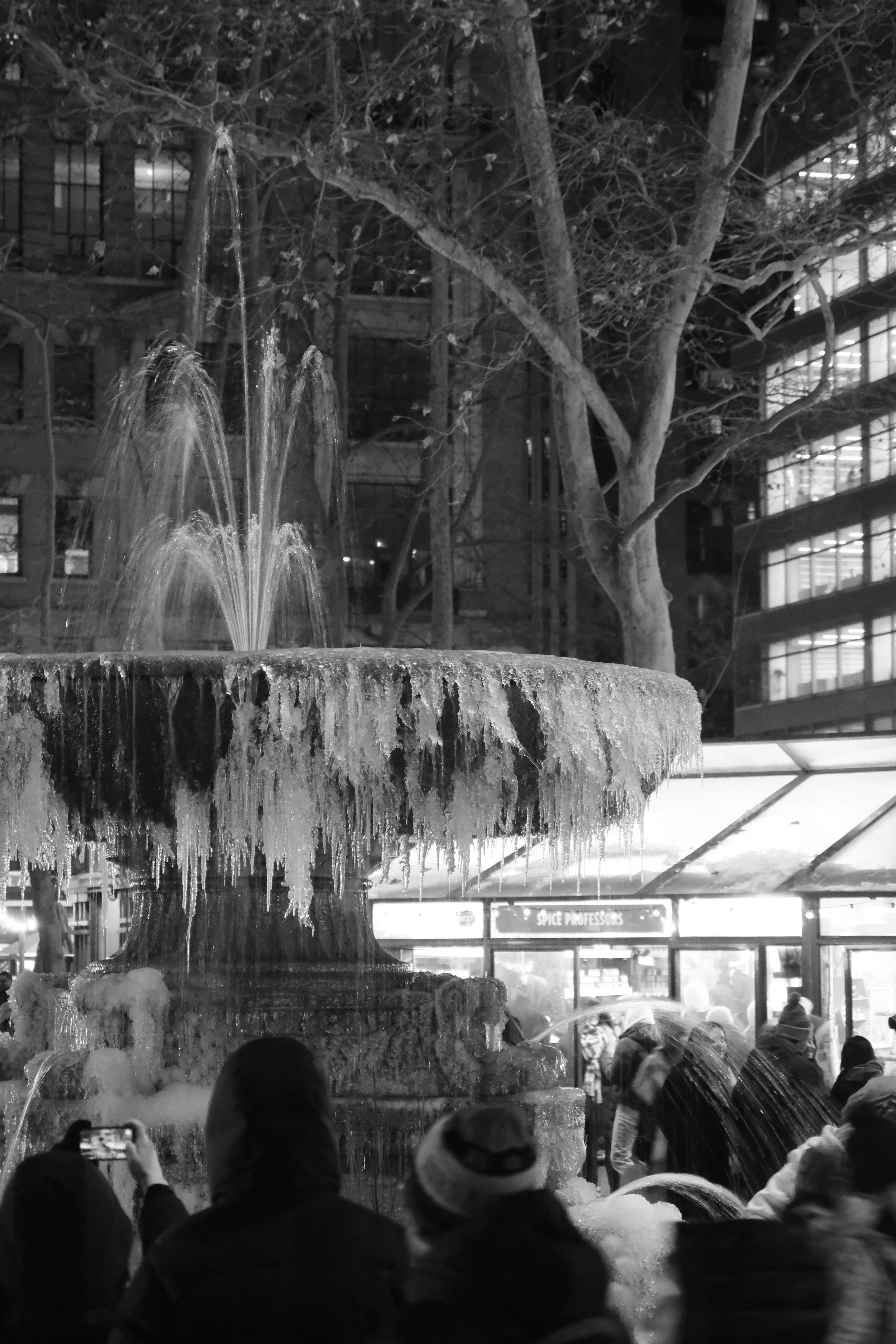 A nighttime scene of a crowd gathered around a large, icy fountain with water cascading over ice formations. In the background, there are modern glass buildings and a leafless tree.