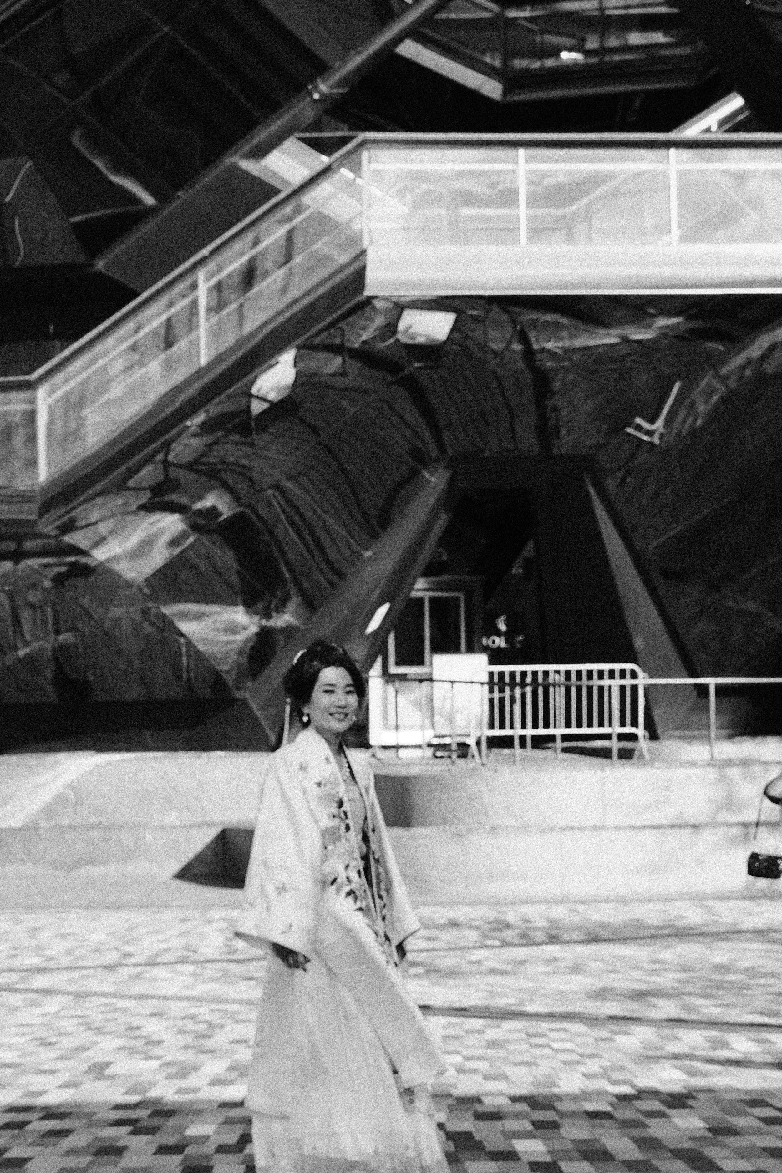 A woman dressed in traditional Japanese attire standing outdoors in front of a modern building with angular architecture and reflective surfaces.