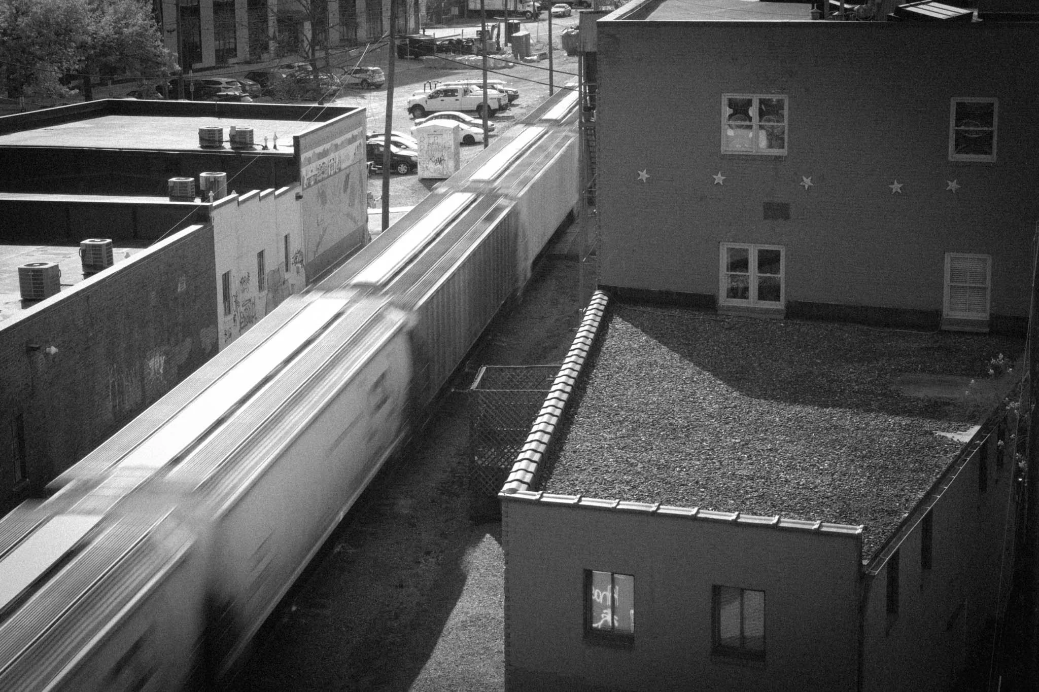 A black and white photo of a moving train passing between buildings in an urban area, with parked cars and a street visible in the background.
