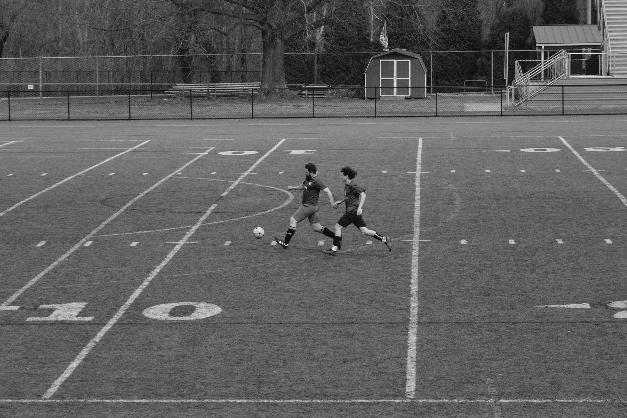 Two boys playing soccer on a school turf field with markings and goal post in the background, holding hands as they chase the ball.