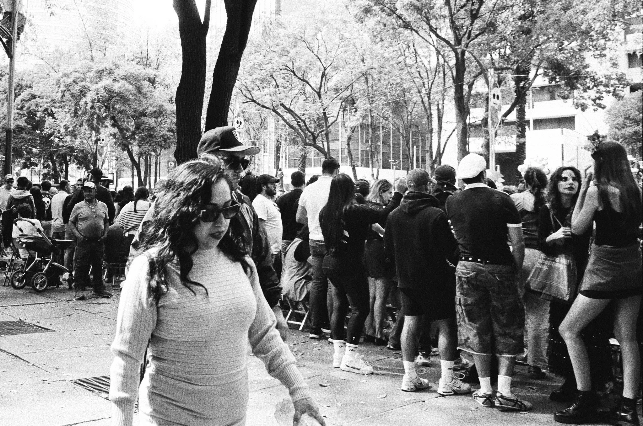 A black-and-white photo of a crowd of people outdoors, some standing and some seated, with trees and buildings in the background.