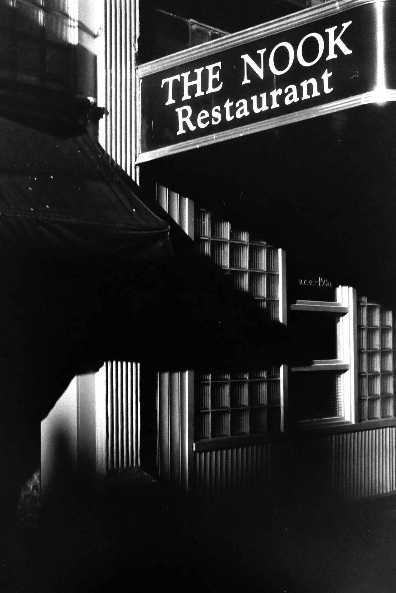 A black-and-white photo of the exterior of a restaurant named 'The Nook.' The sign is prominent, and there are bookshelves and architectural details visible in the background.