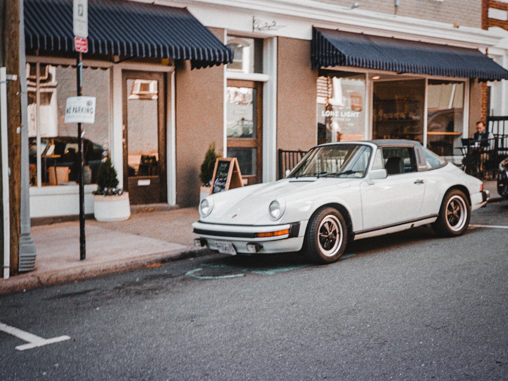 A vintage white Porsche 911 parked in front of a storefront with blue awnings, a sidewalk chalkboard sign, and outdoor seating, with a person sitting at a table in the background.