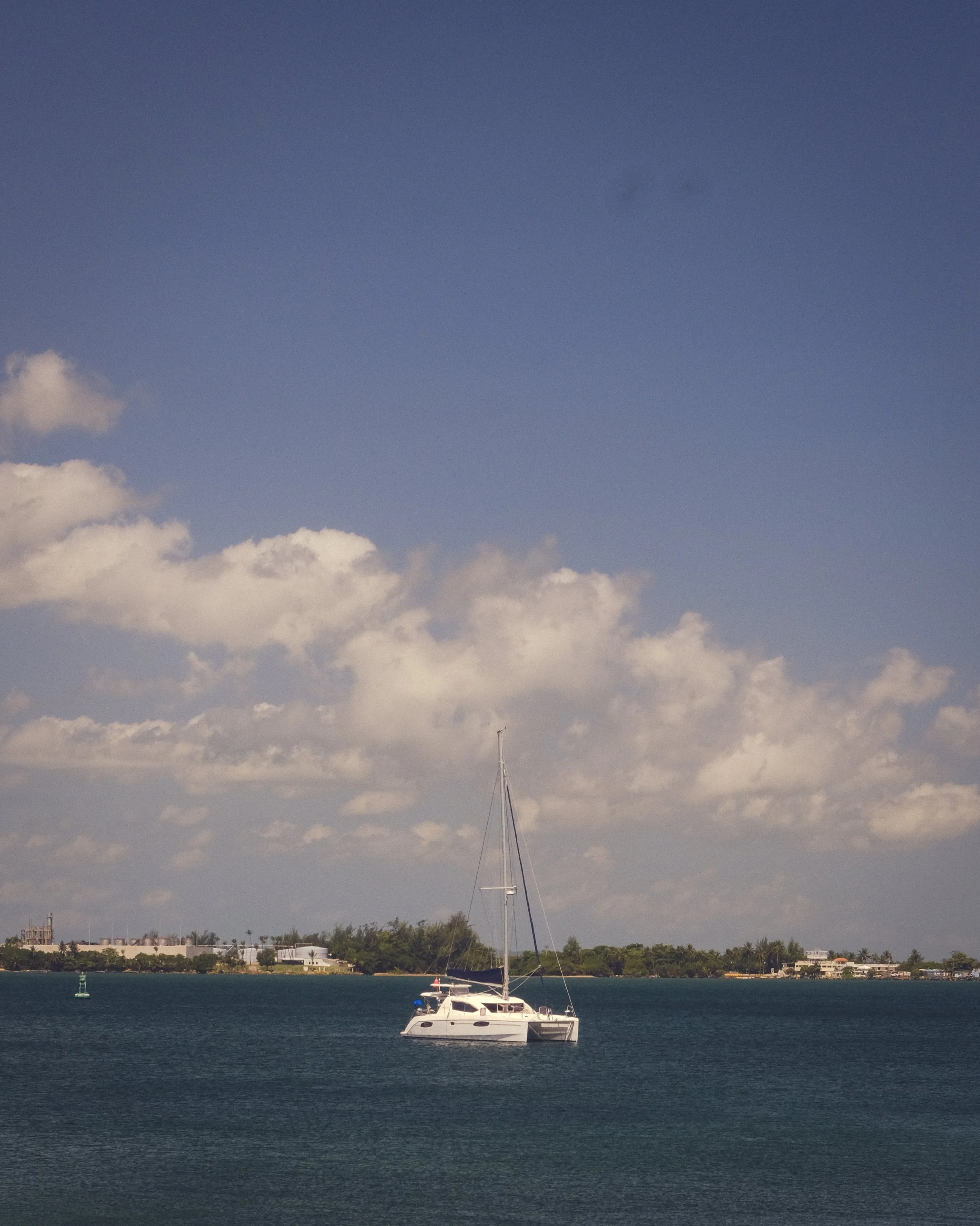 A white sailboat with a tall mast floating on a body of water under a partly cloudy sky, with land and buildings visible in the background.