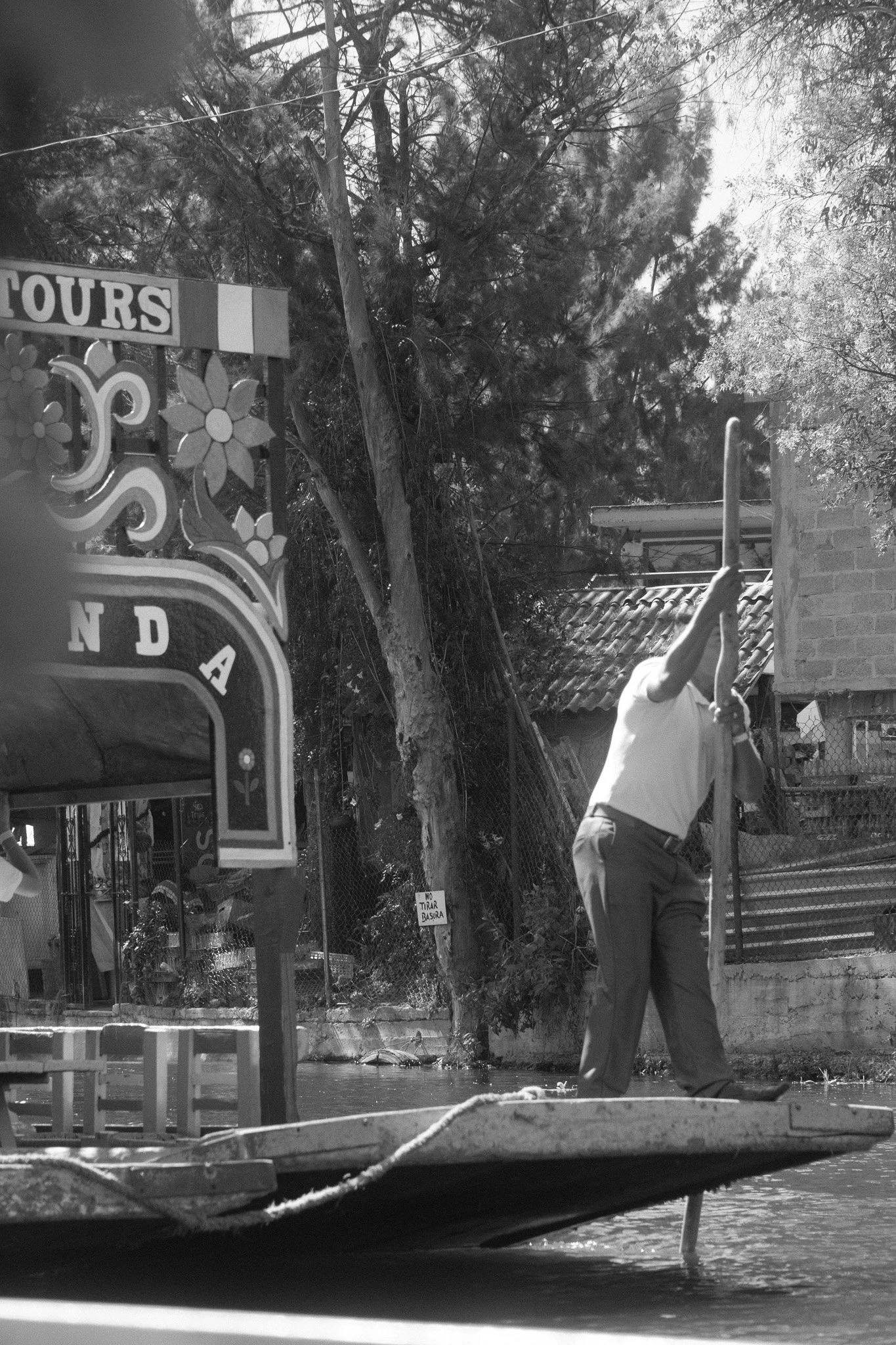 A person in a white shirt standing on a boat with a pole, on a canal with trees and buildings in the background.